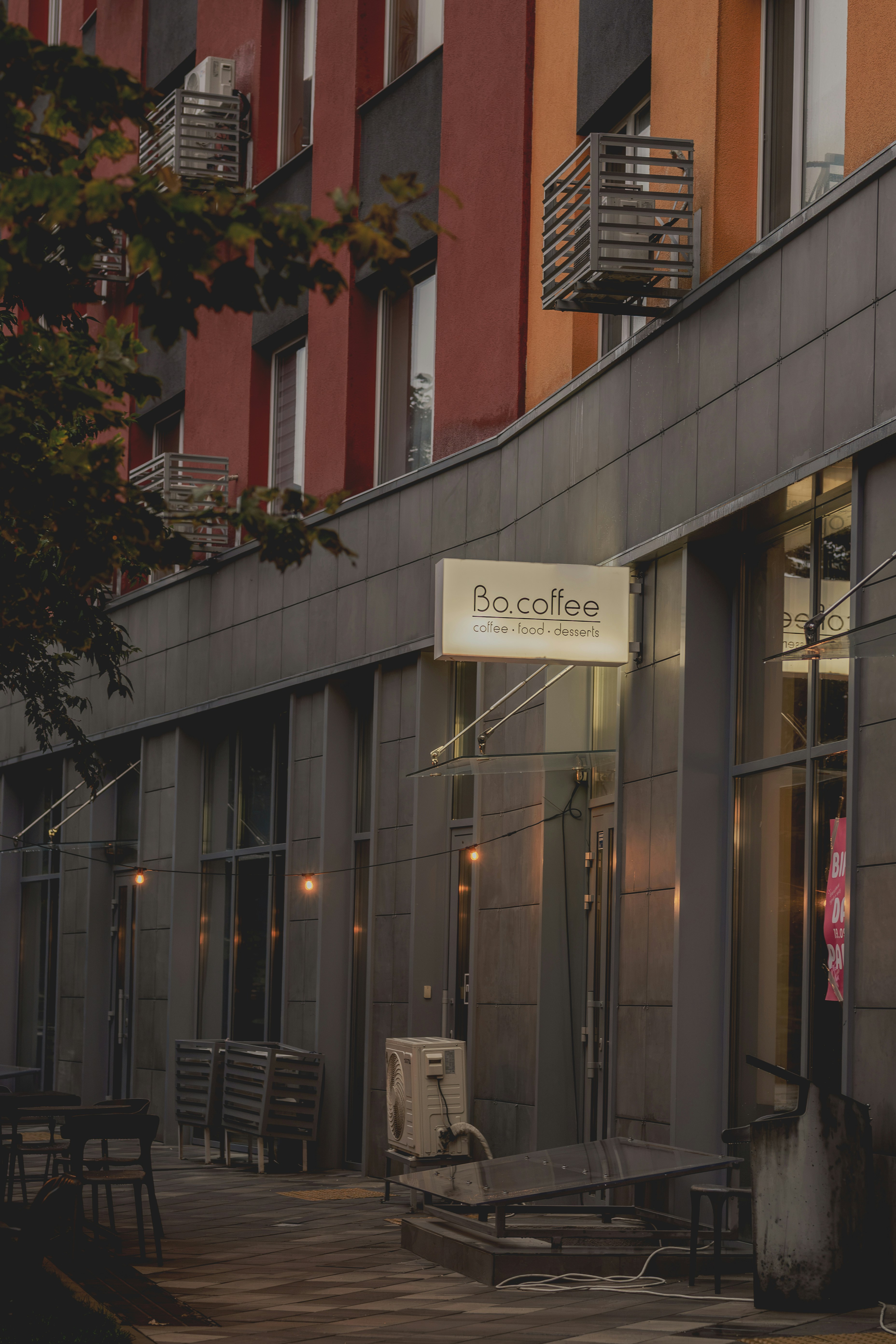 Un escaparate de un café con un cartel luminoso que dice Bo.coffee sobre una acera con terrazas al aire libre