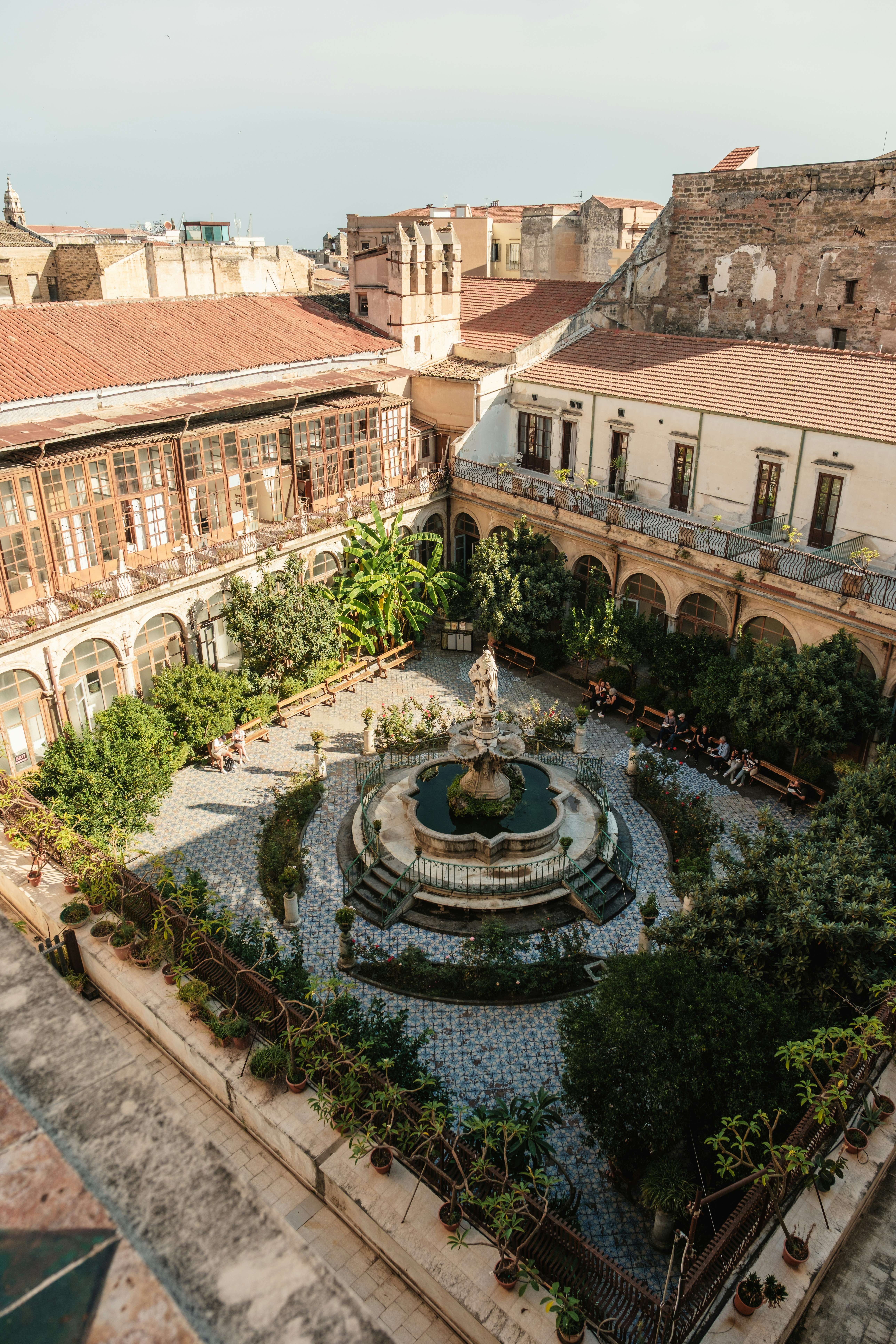 An aerial view of a courtyard with a central fountain surrounded by trees and benches in Palermo