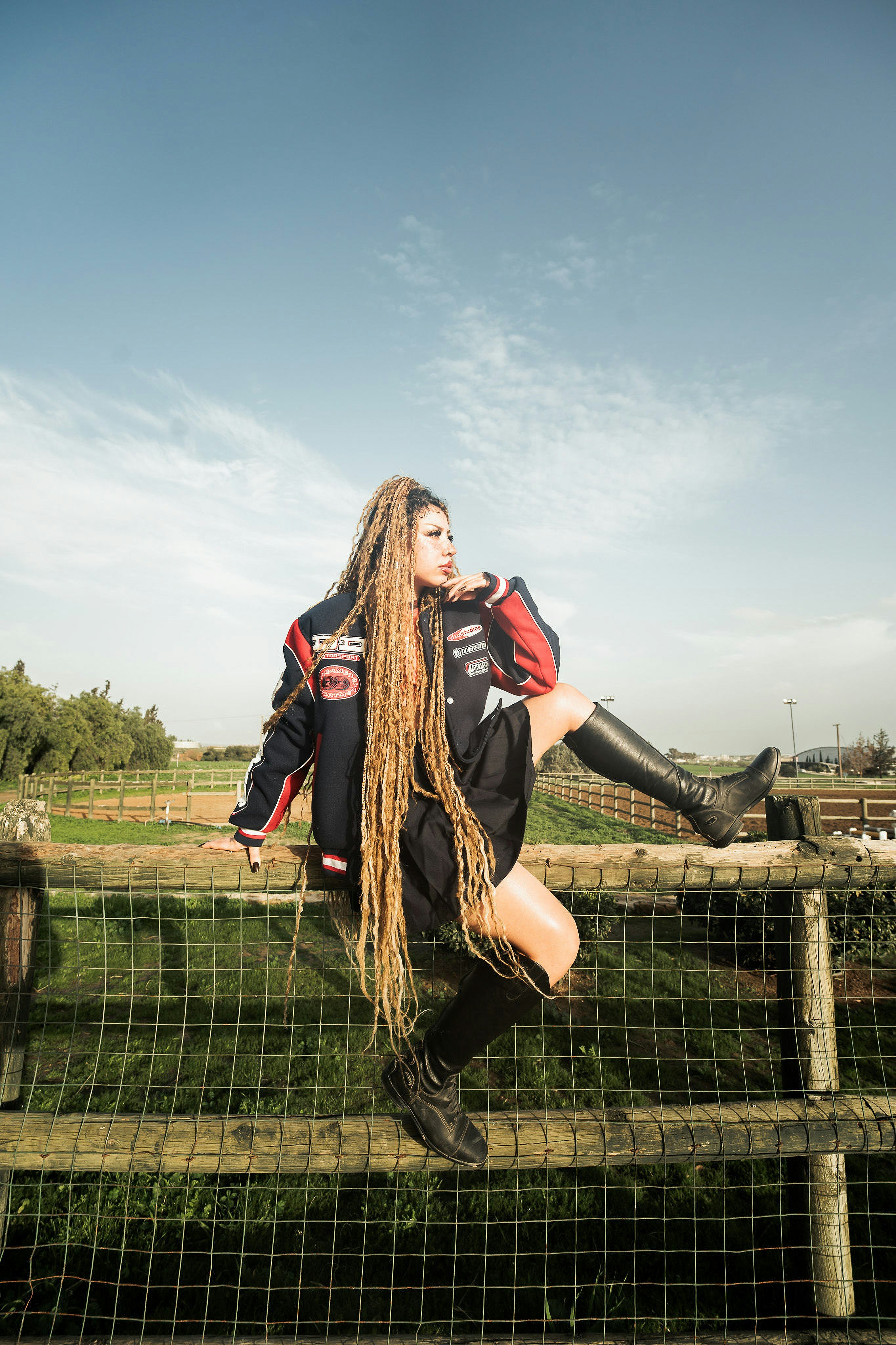 A person with long blonde dreadlocks wearing a varsity jacket and boots on a wooden fence