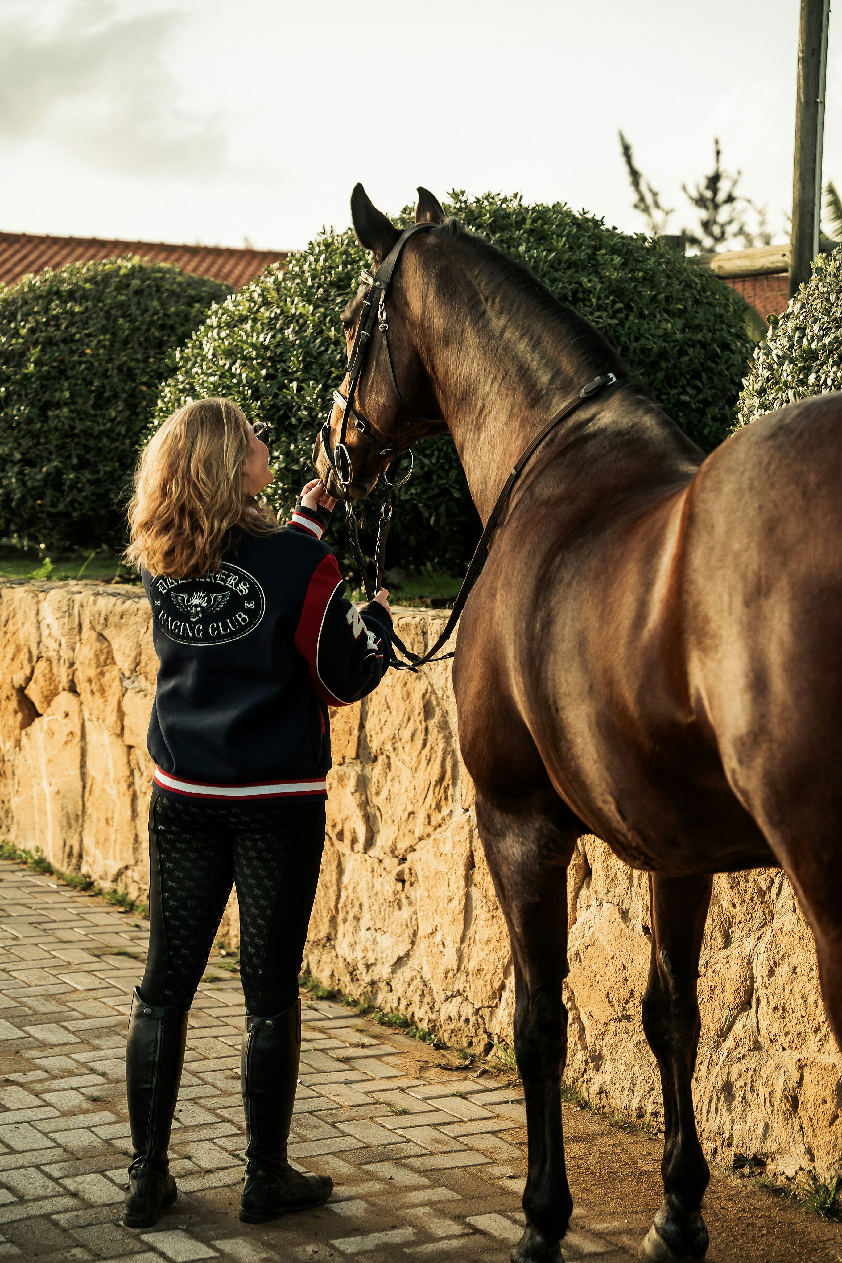 A woman in a varsity jacket and riding boots interacting with a brown horse outdoors