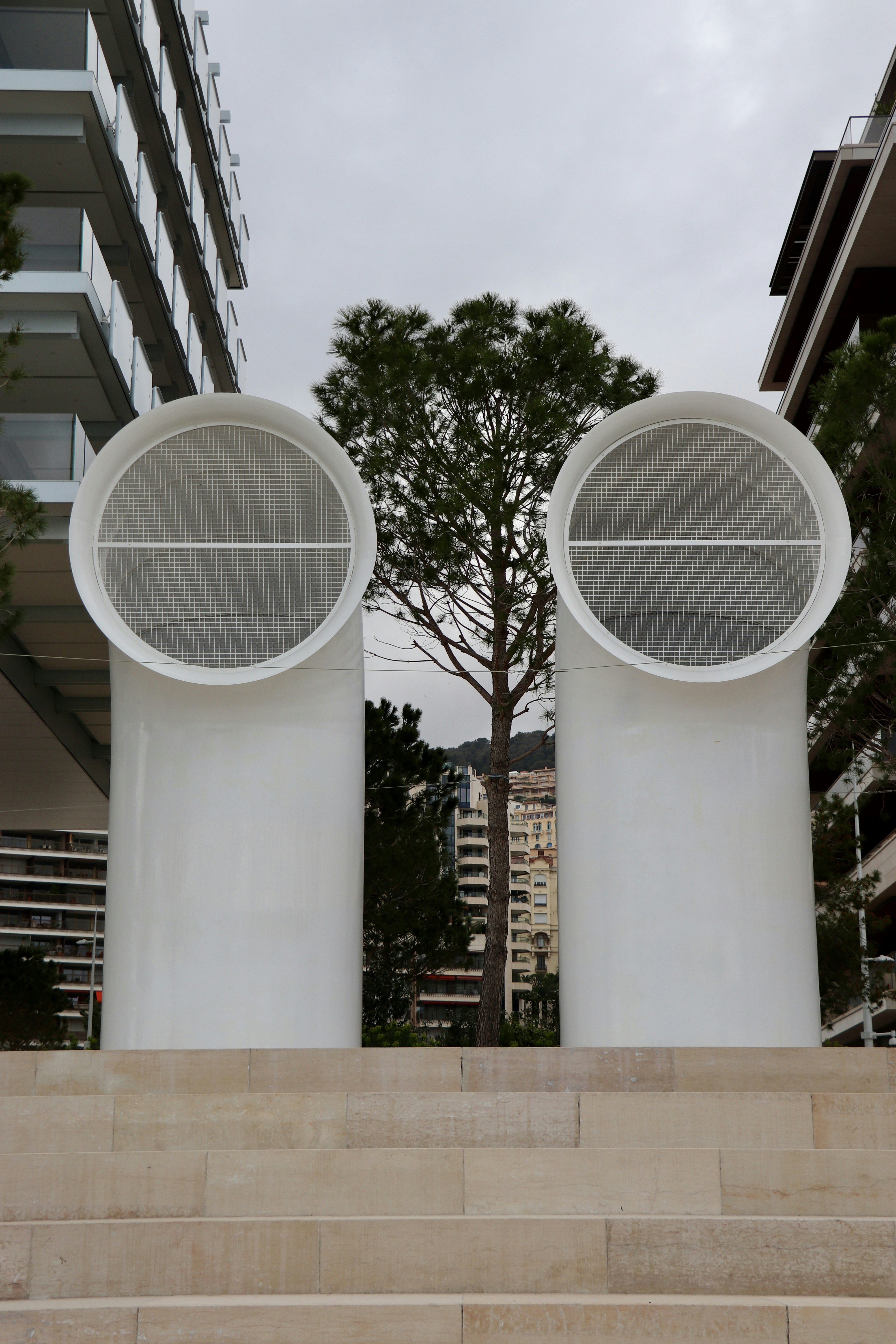 Two large white ventilation pipes with circular grilles standing on stone steps before a pine tree
