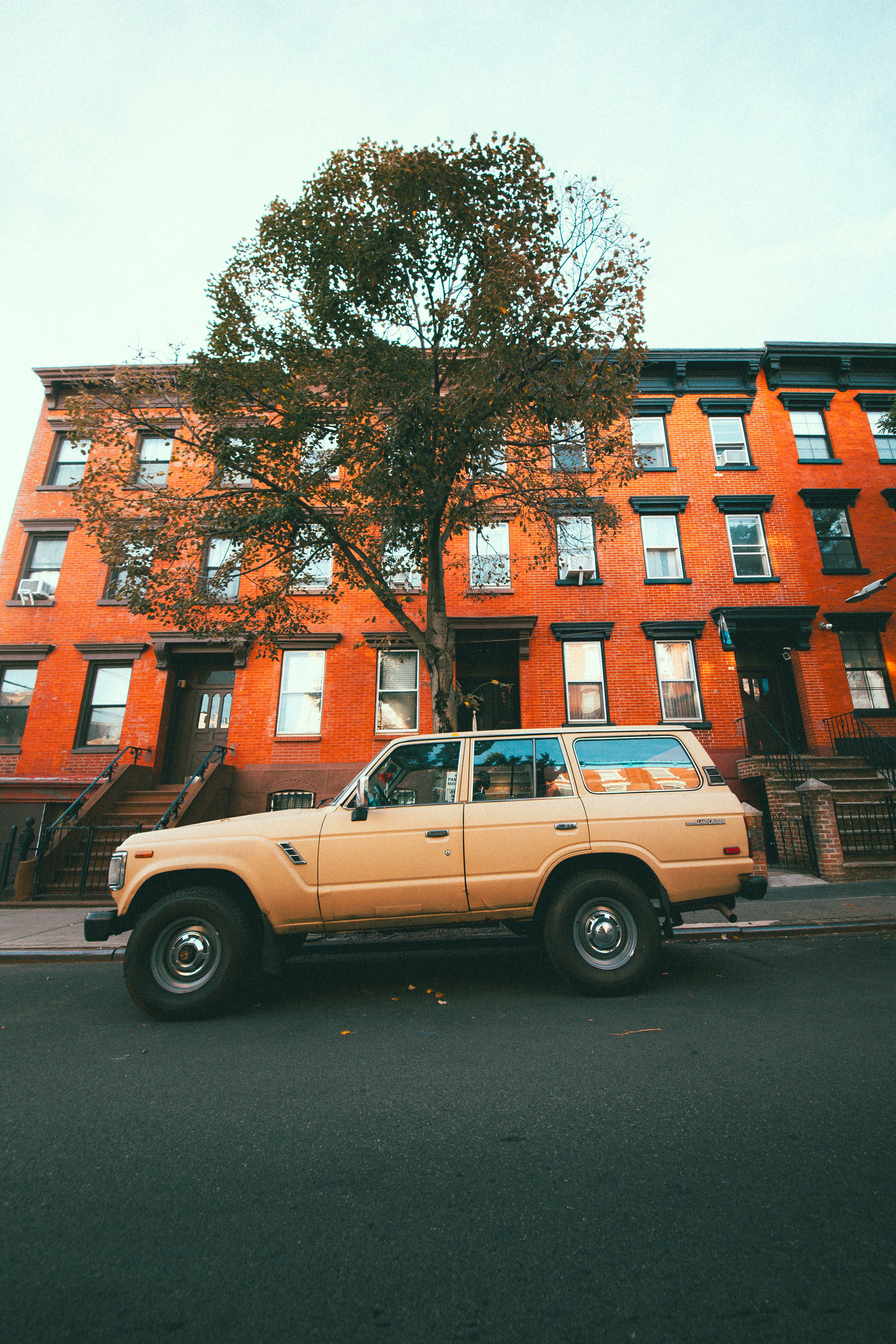 A tan vintage SUV parked on a street in front of orange brick row houses in New York