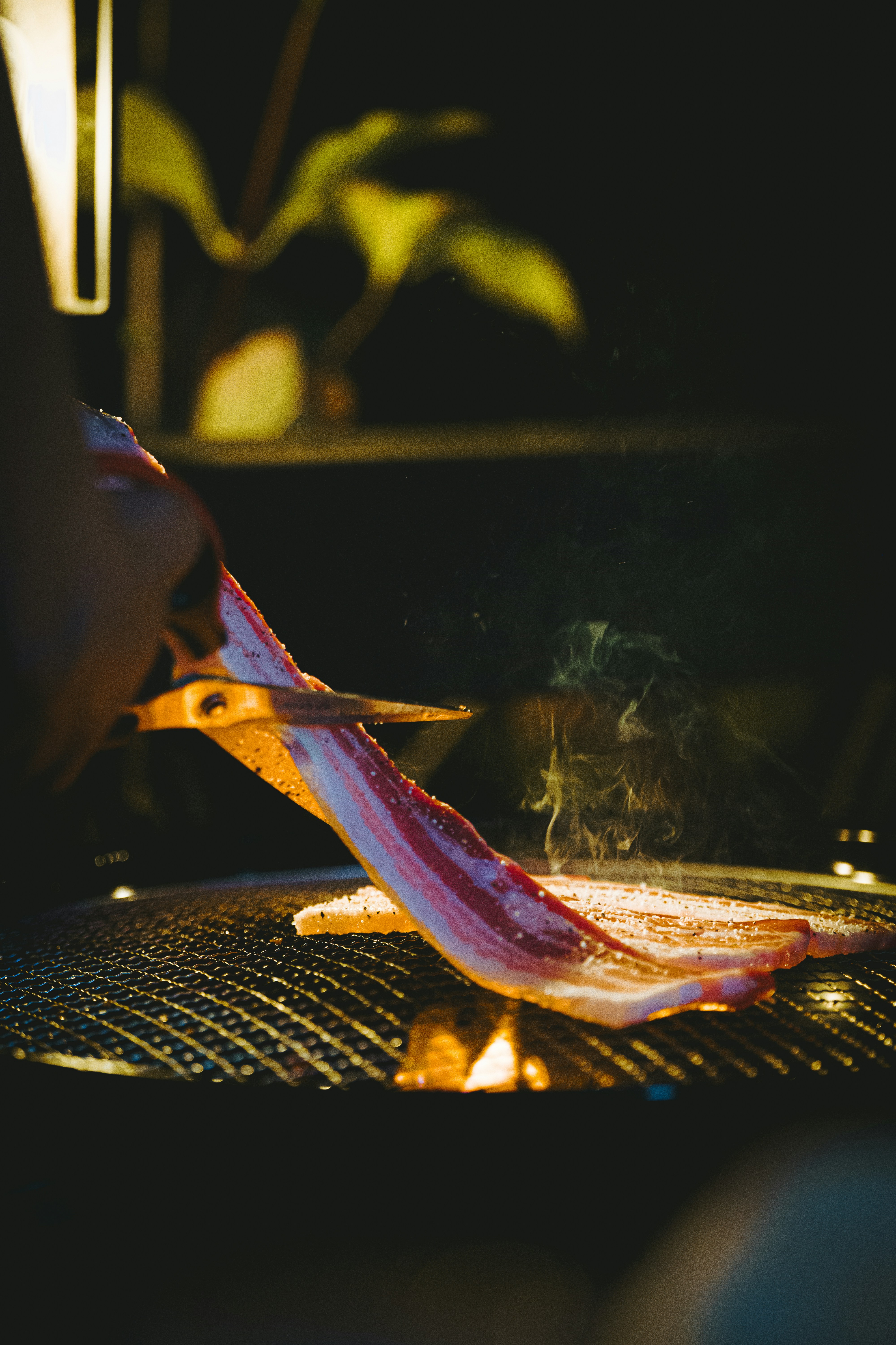 A person using scissors to cut a strip of raw bacon on a hot grill