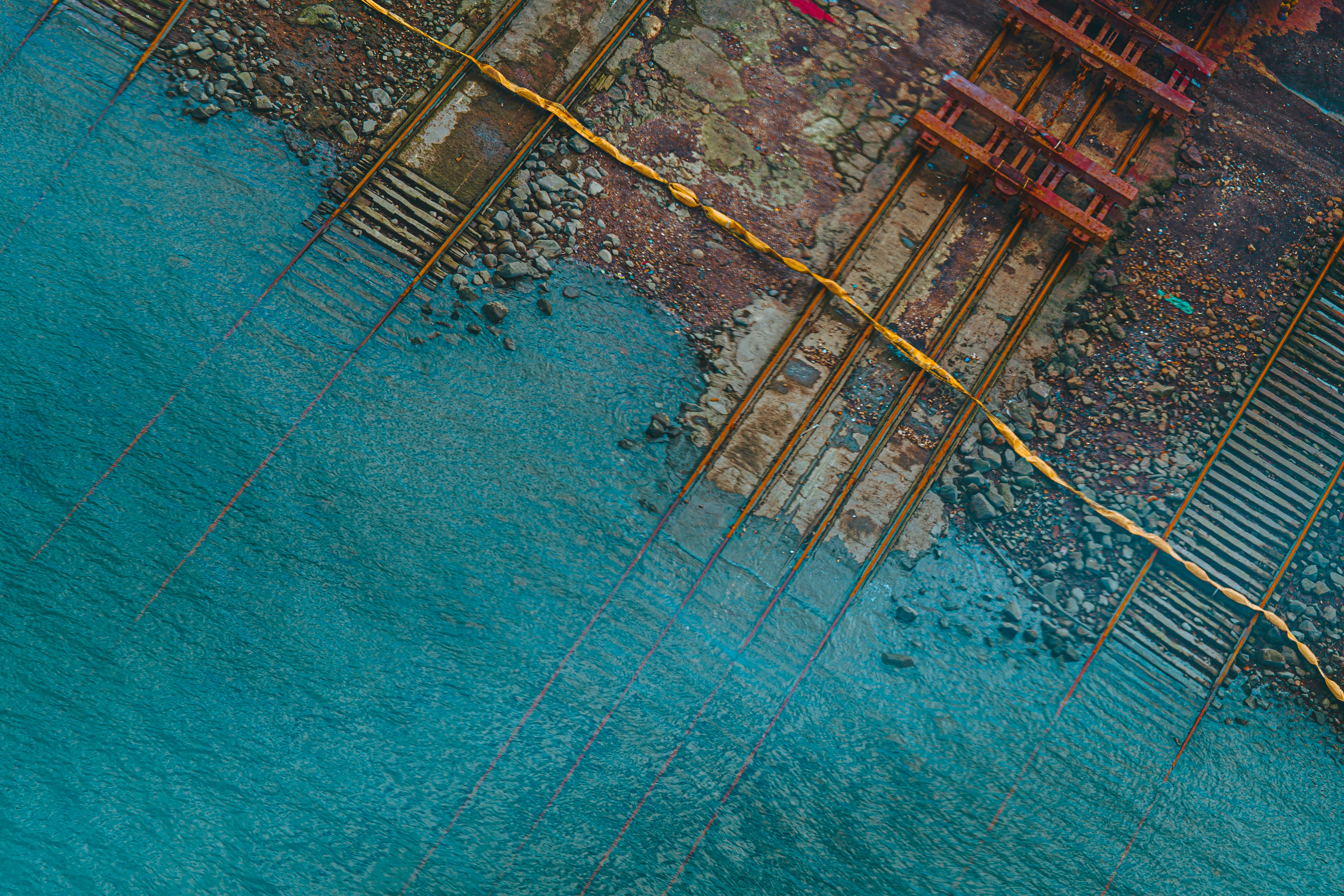 An aerial view of rusted railway tracks extending into turquoise water from a rocky shore