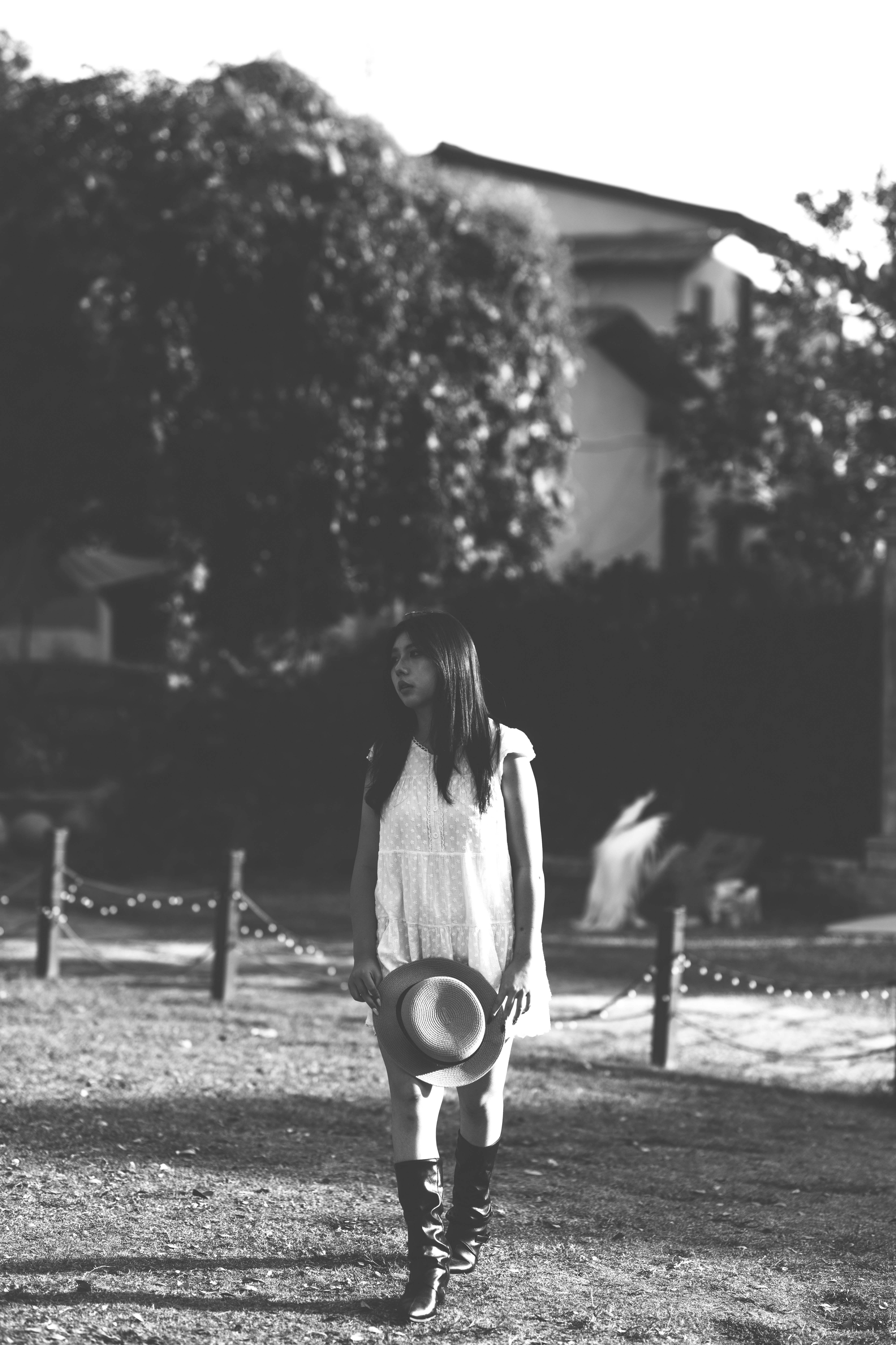 A woman in a white dress and boots holding a hat in a sunlit garden