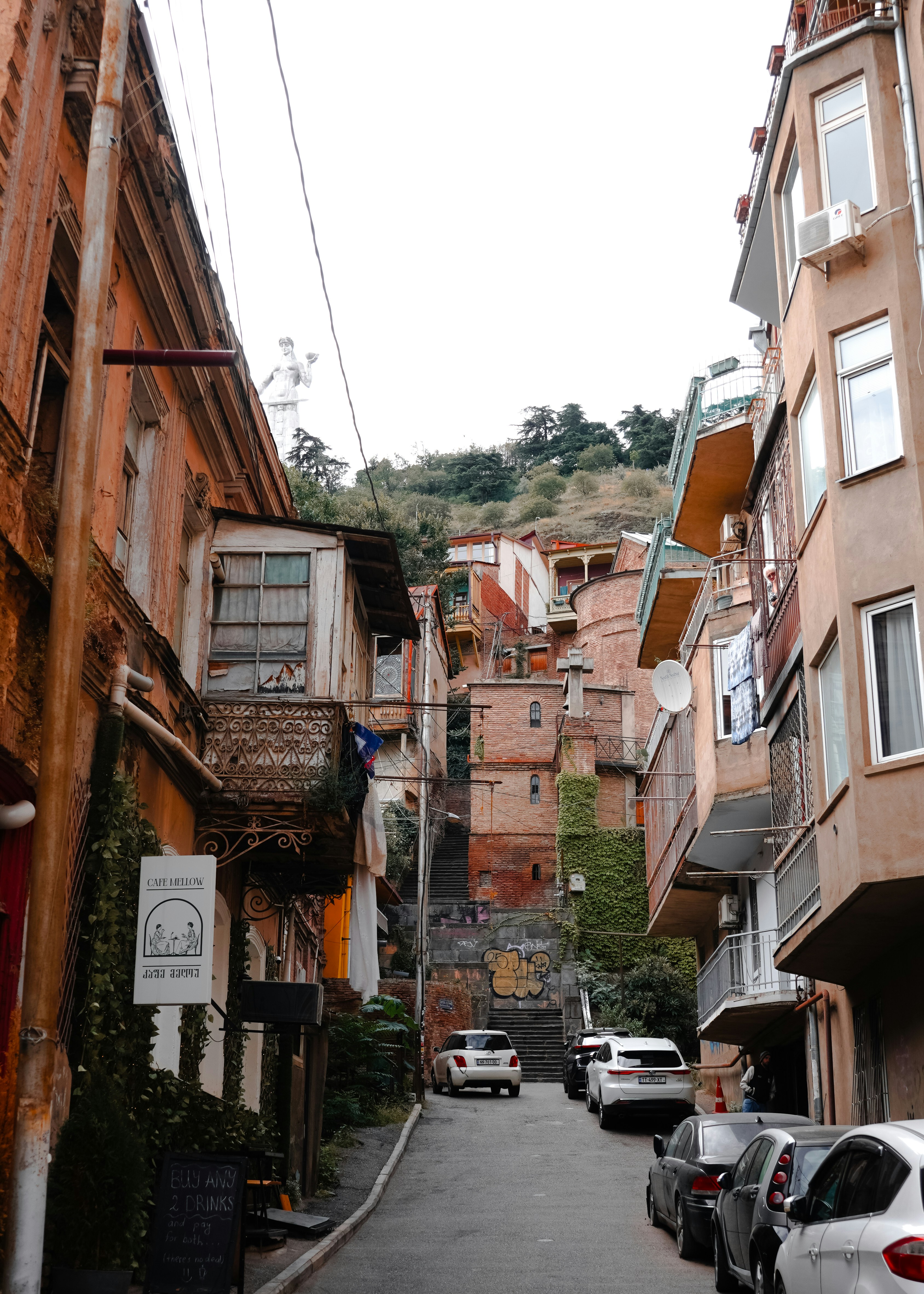 A narrow street in Tbilisi with historic buildings, parked cars, and the Kartlis Deda statue on the hill