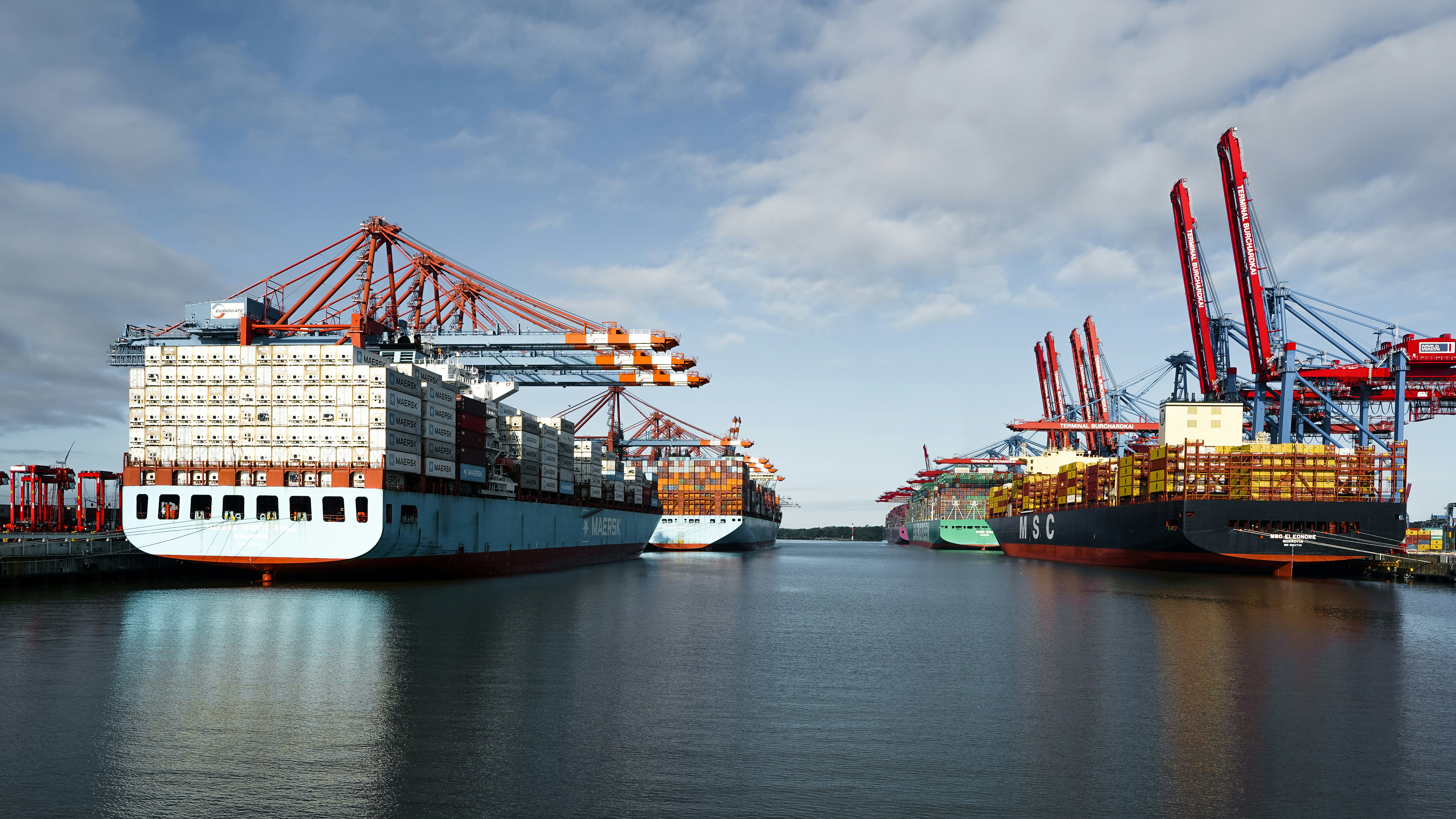 Large container ships docked at a busy industrial port under a cloudy blue sky