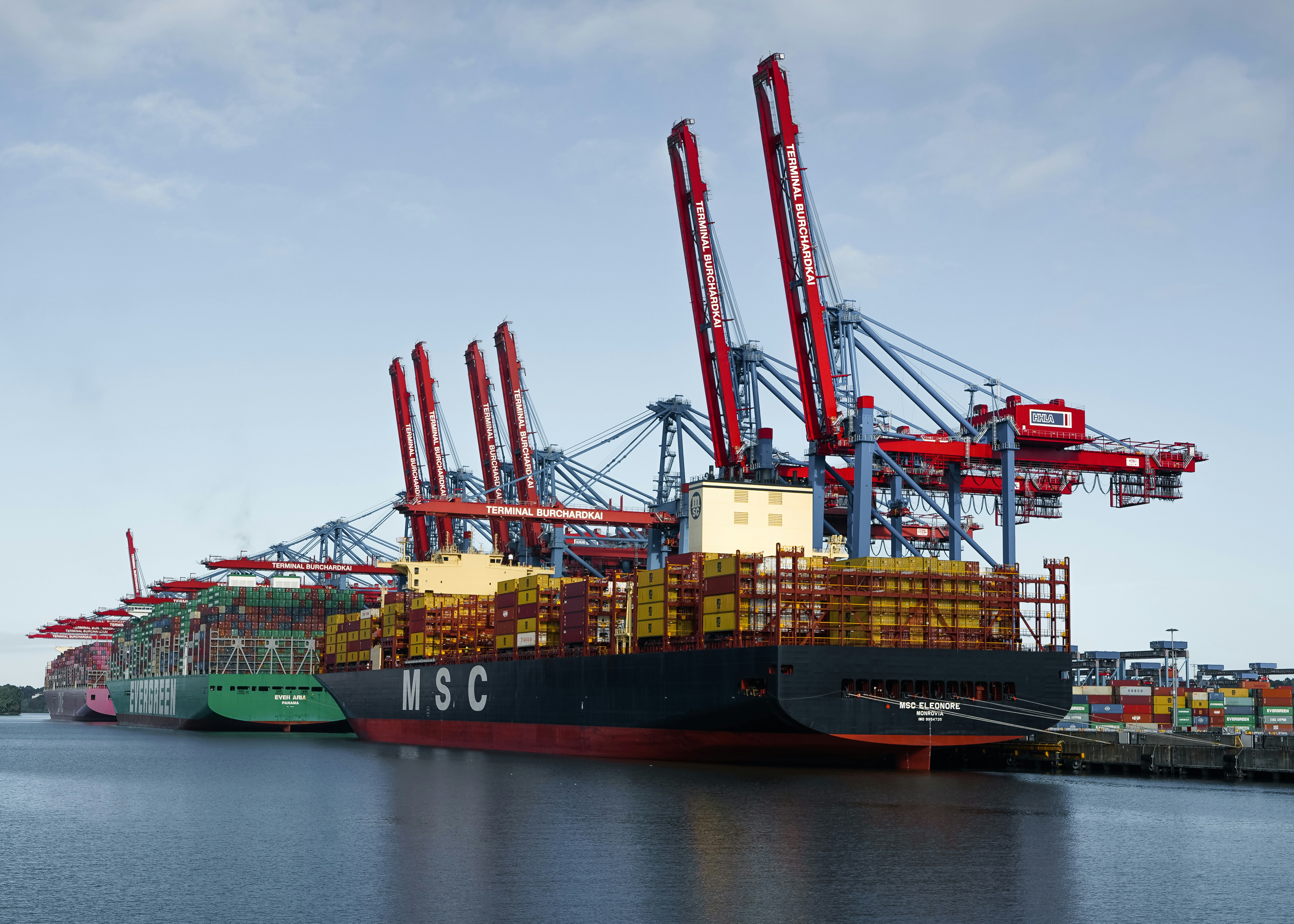 Large container ships docked at a terminal with red cranes under a clear sky