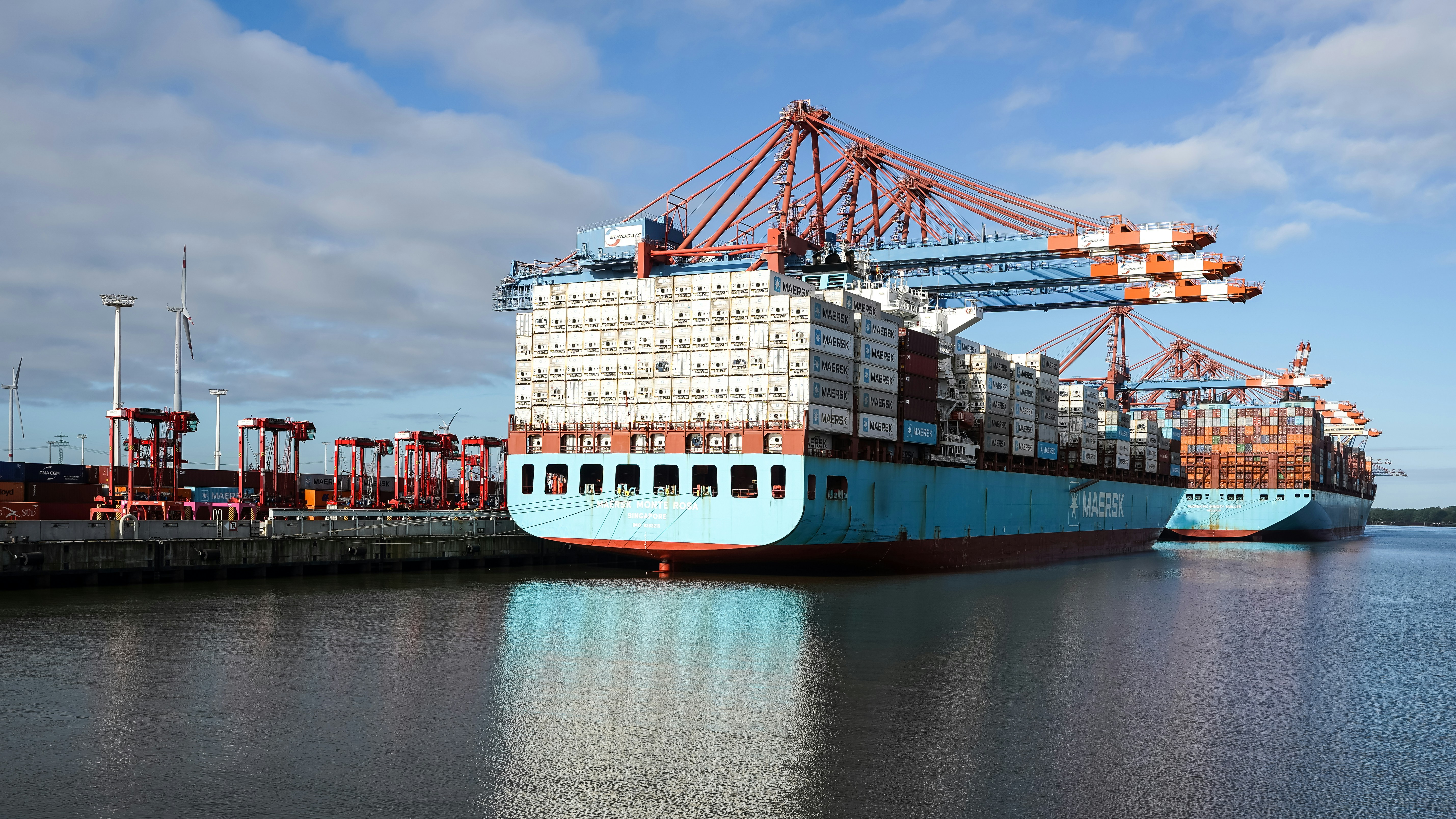 Two large container ships docked at a port with red cranes under a blue sky