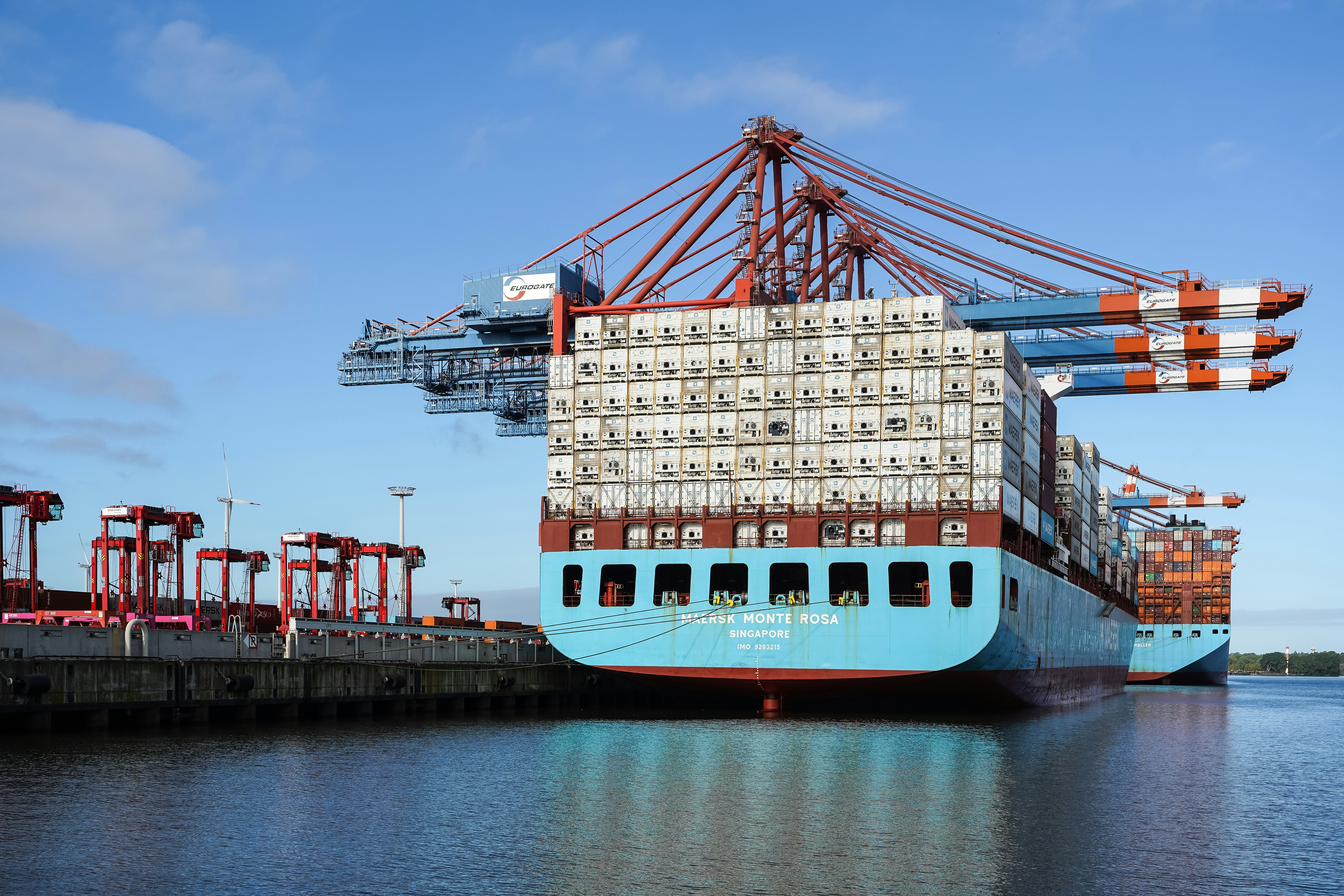 A large container ship docked at a port with red cranes under a blue sky