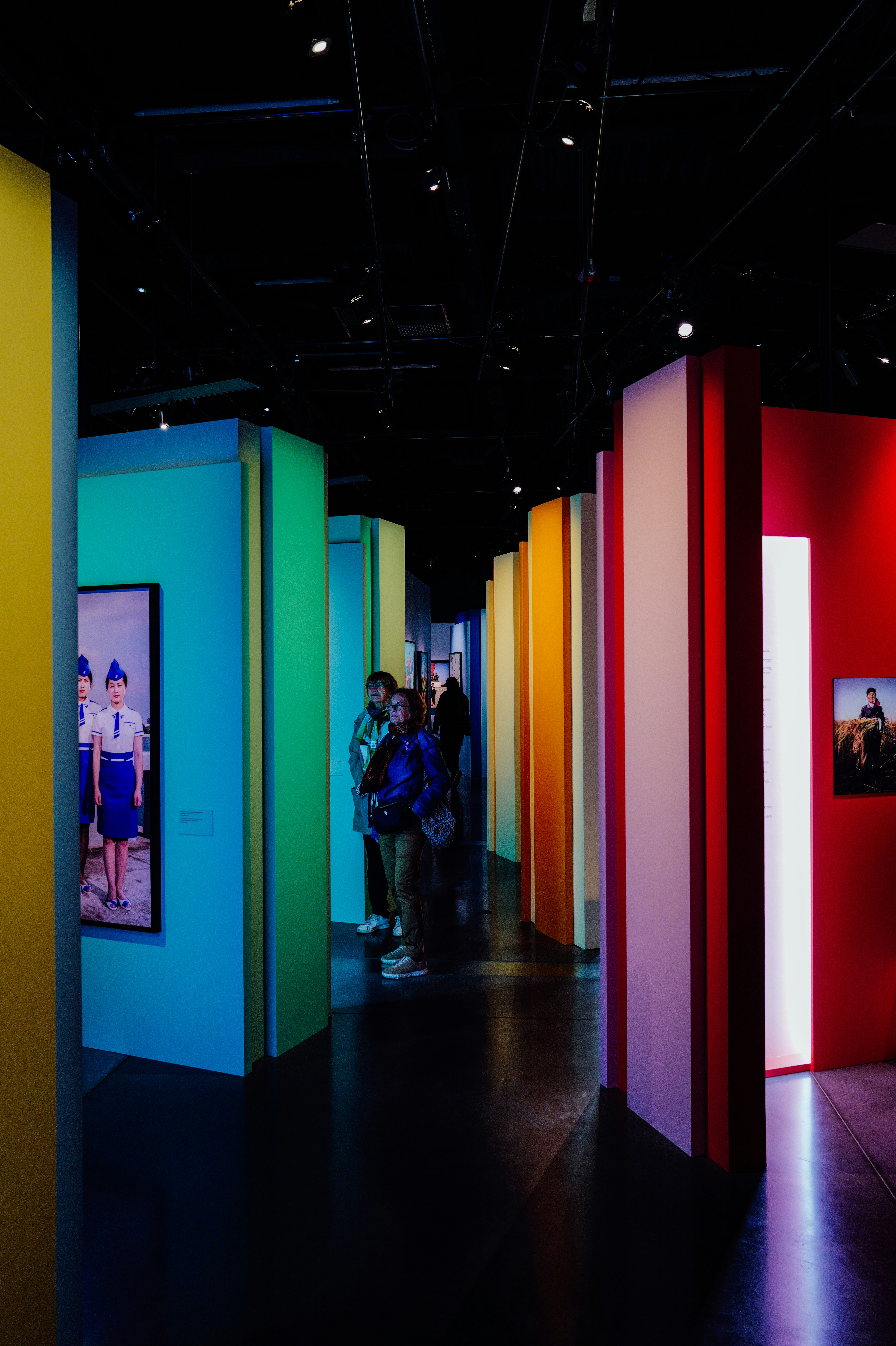 Deux adultes traversant une galerie avec des cloisons colorées et des photographies encadrées aux murs