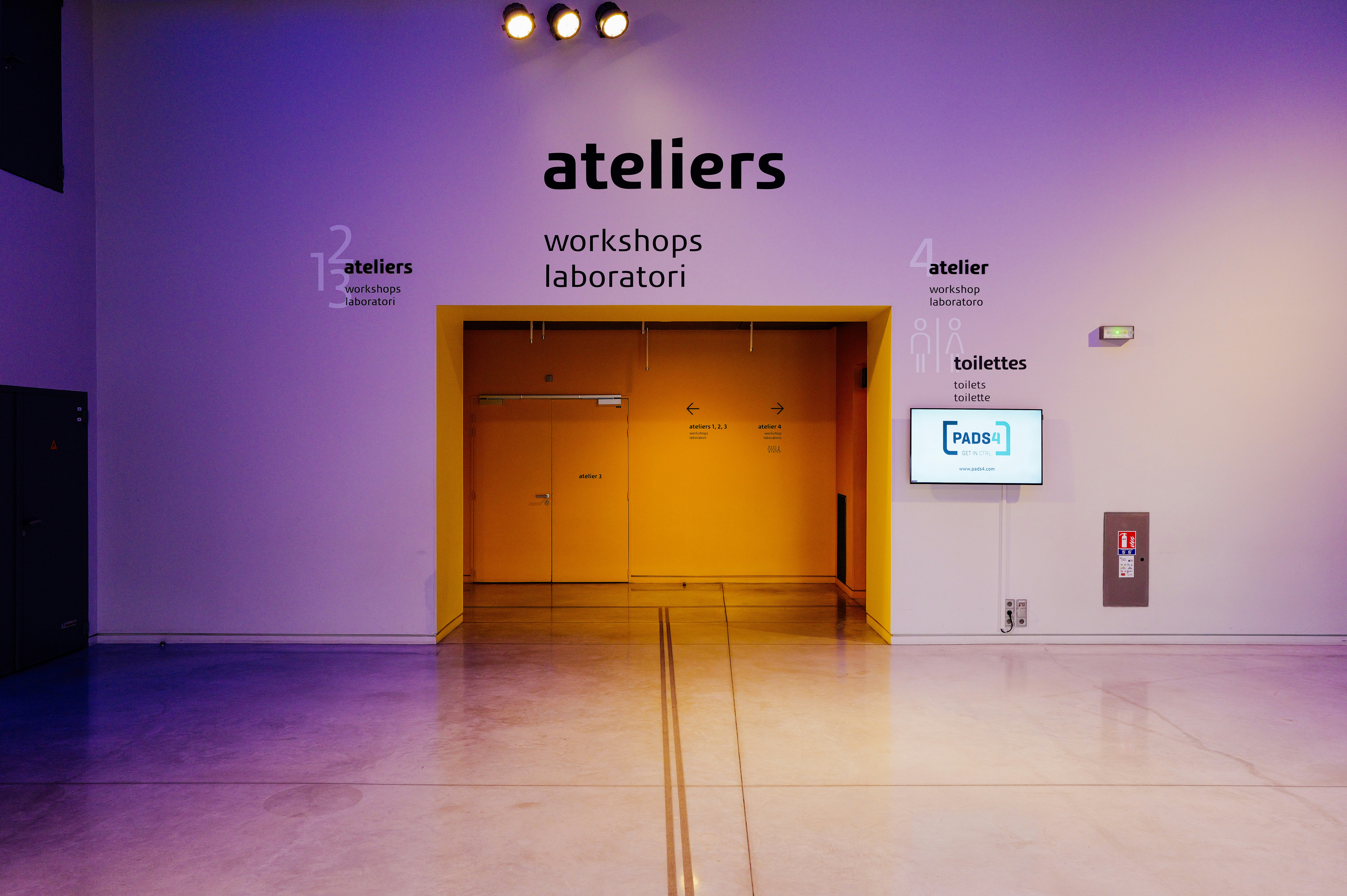 Une porte jaune dans un mur blanc avec une signalétique indiquant les ateliers et les toilettes