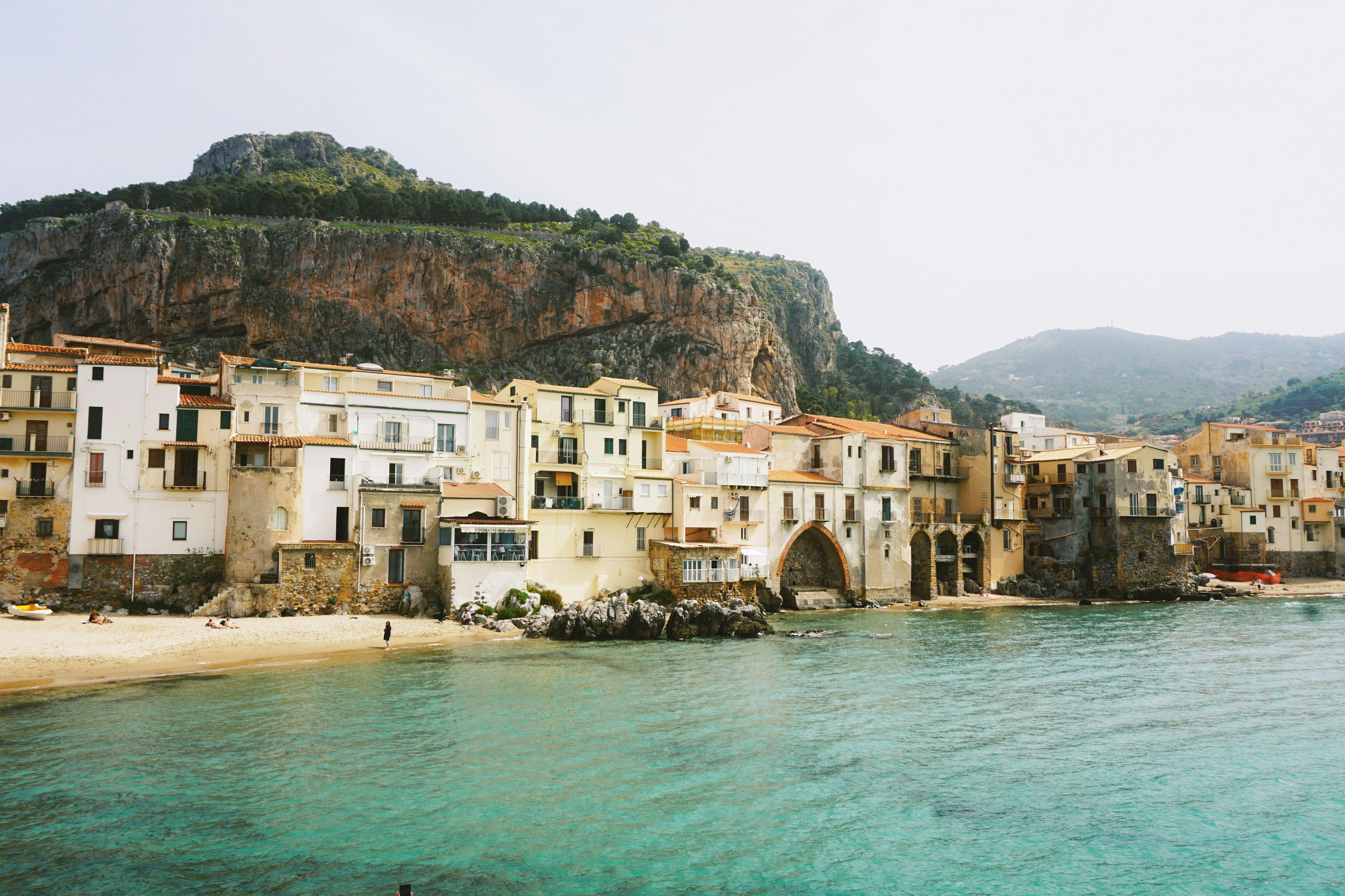 Buildings along the sandy beach of Cefalù beneath a large rocky cliff in Sicily