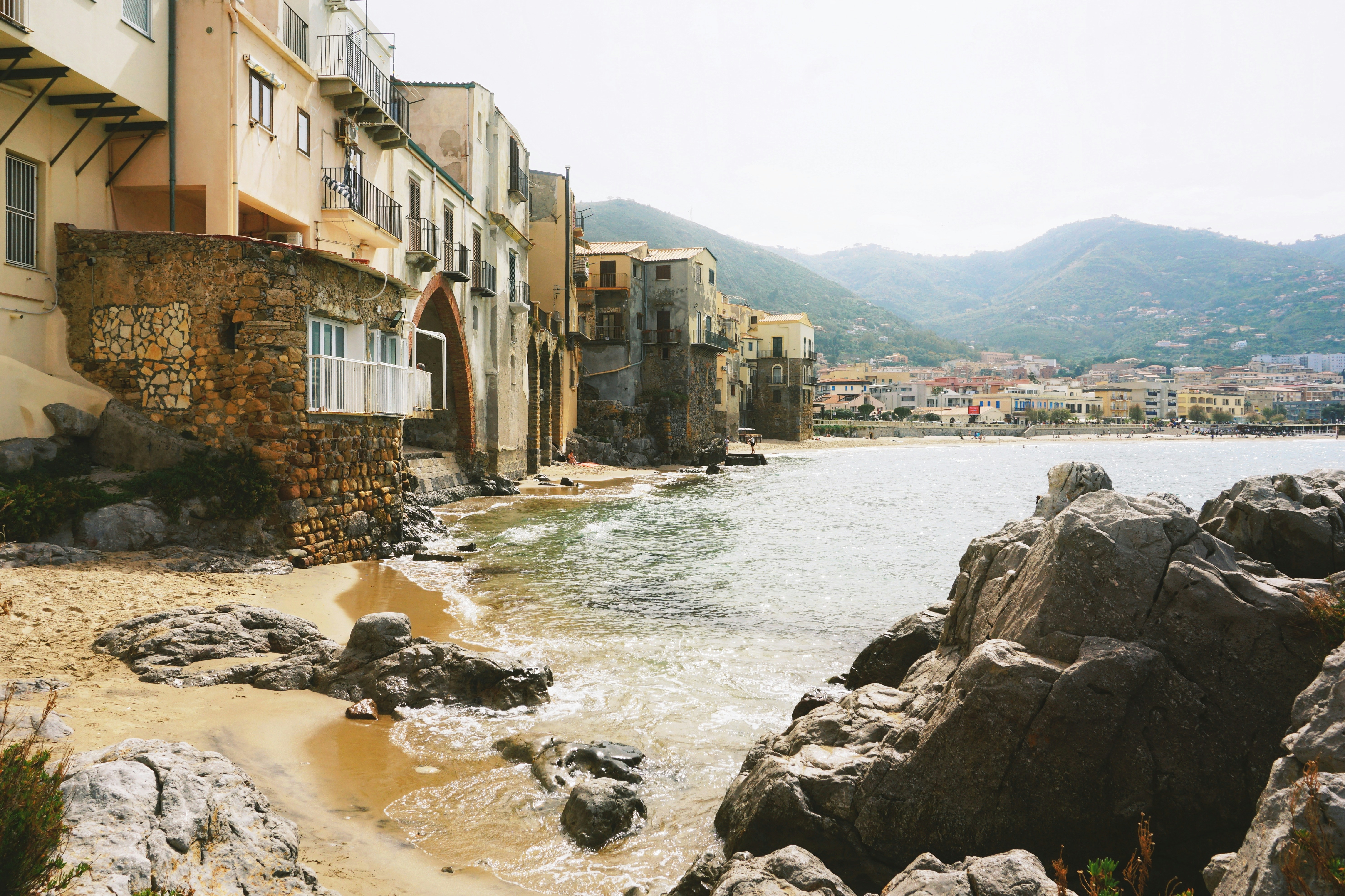 Old stone buildings along a sandy beach with rocky outcrops and distant mountains in Cefalù