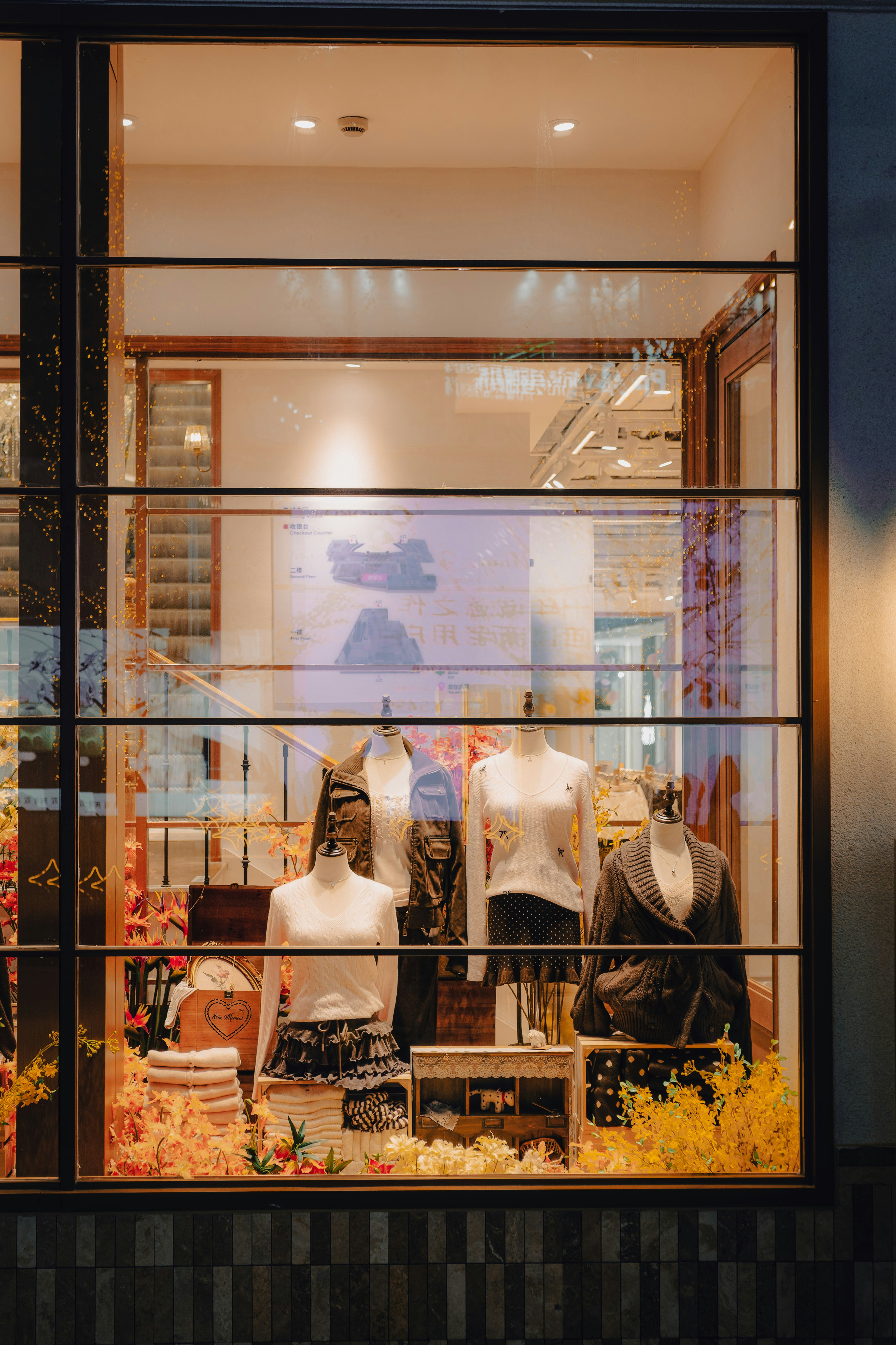 Clothing store window display with mannequins wearing sweaters and skirts surrounded by yellow flowers