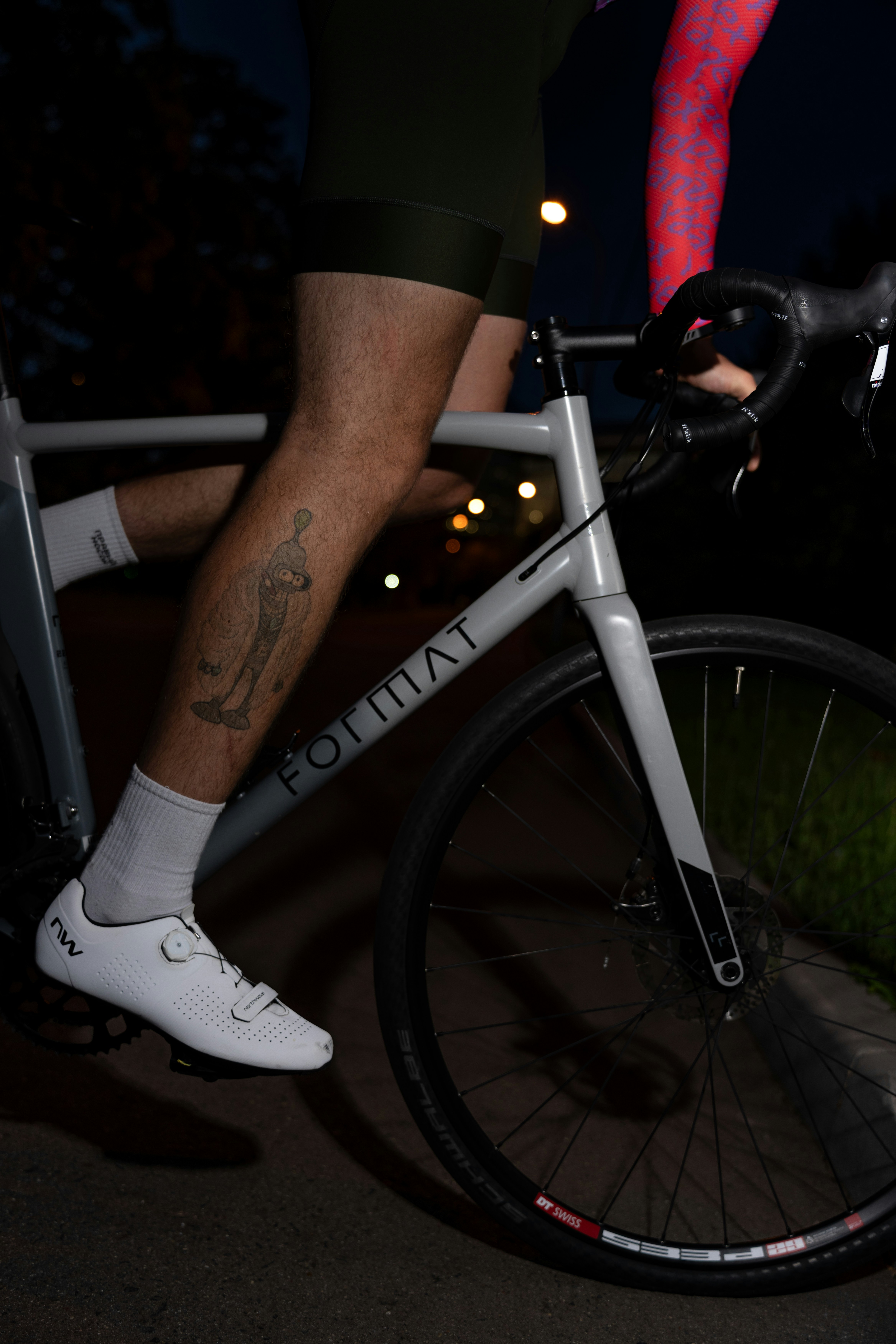 A cyclist with a cartoon tattoo on their leg riding a grey road bike at night