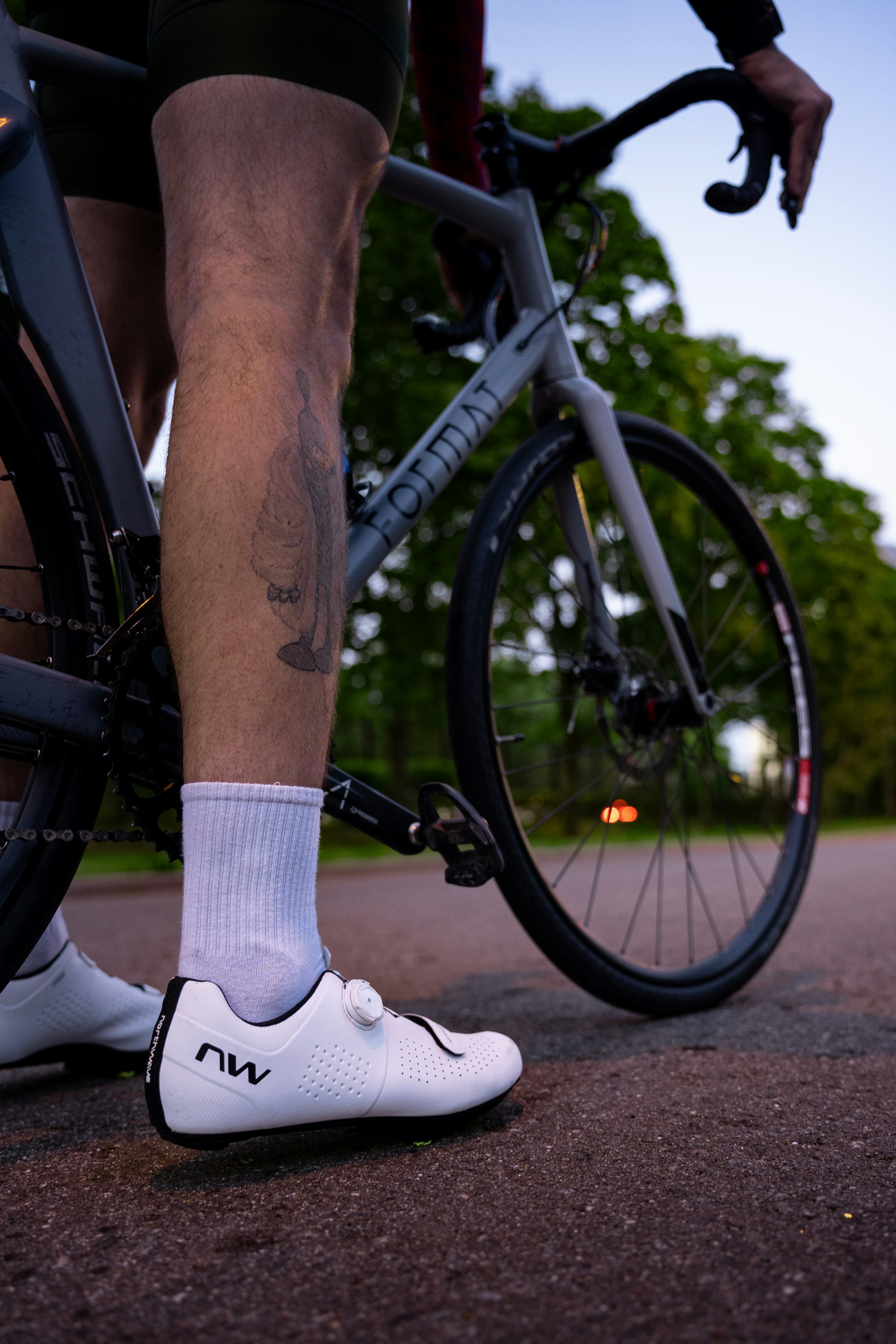 A cyclist with a leg tattoo wearing white cycling shoes on a road bike