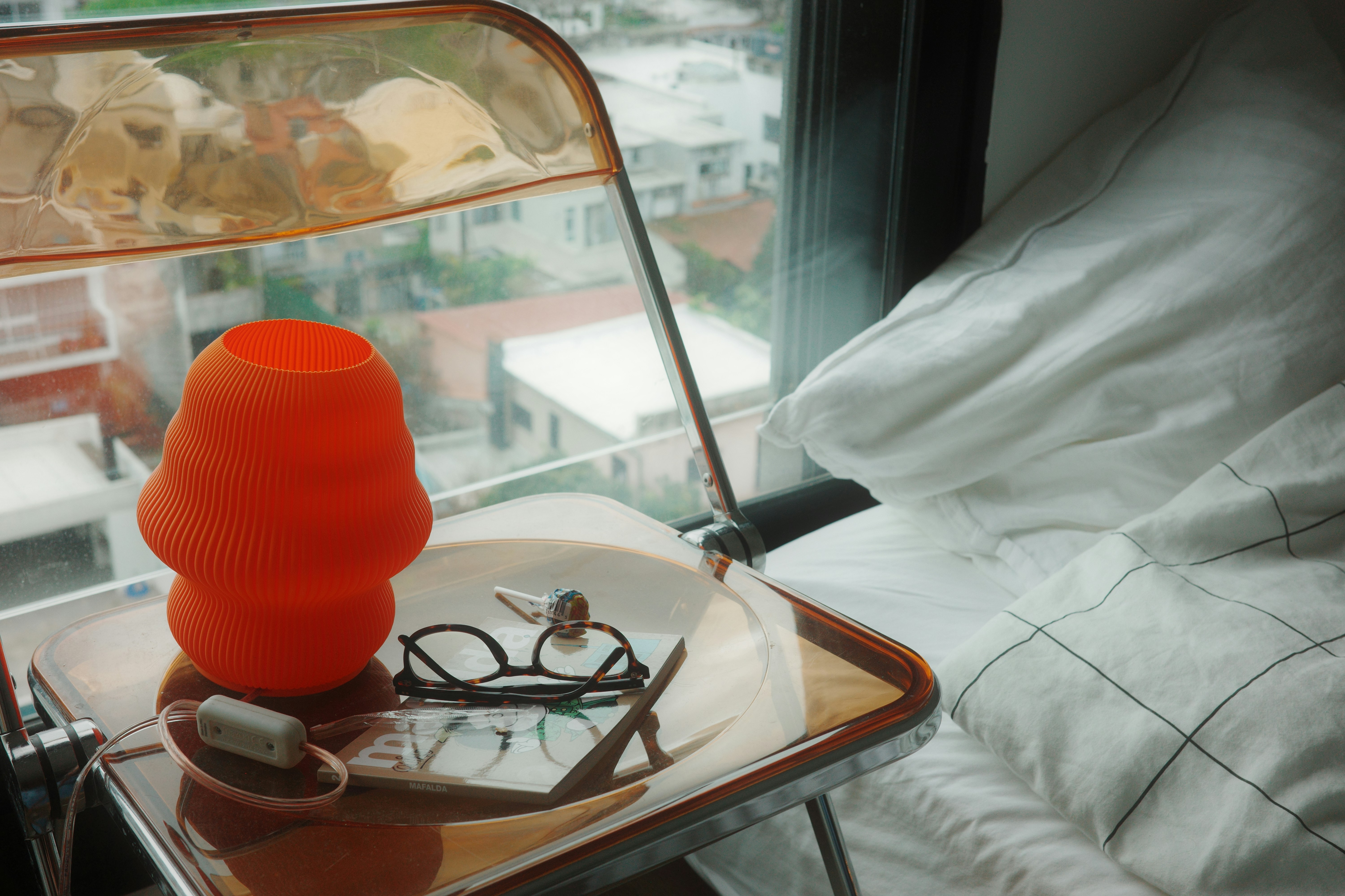 An orange lamp, glasses, and a small book on a transparent side table beside a bed