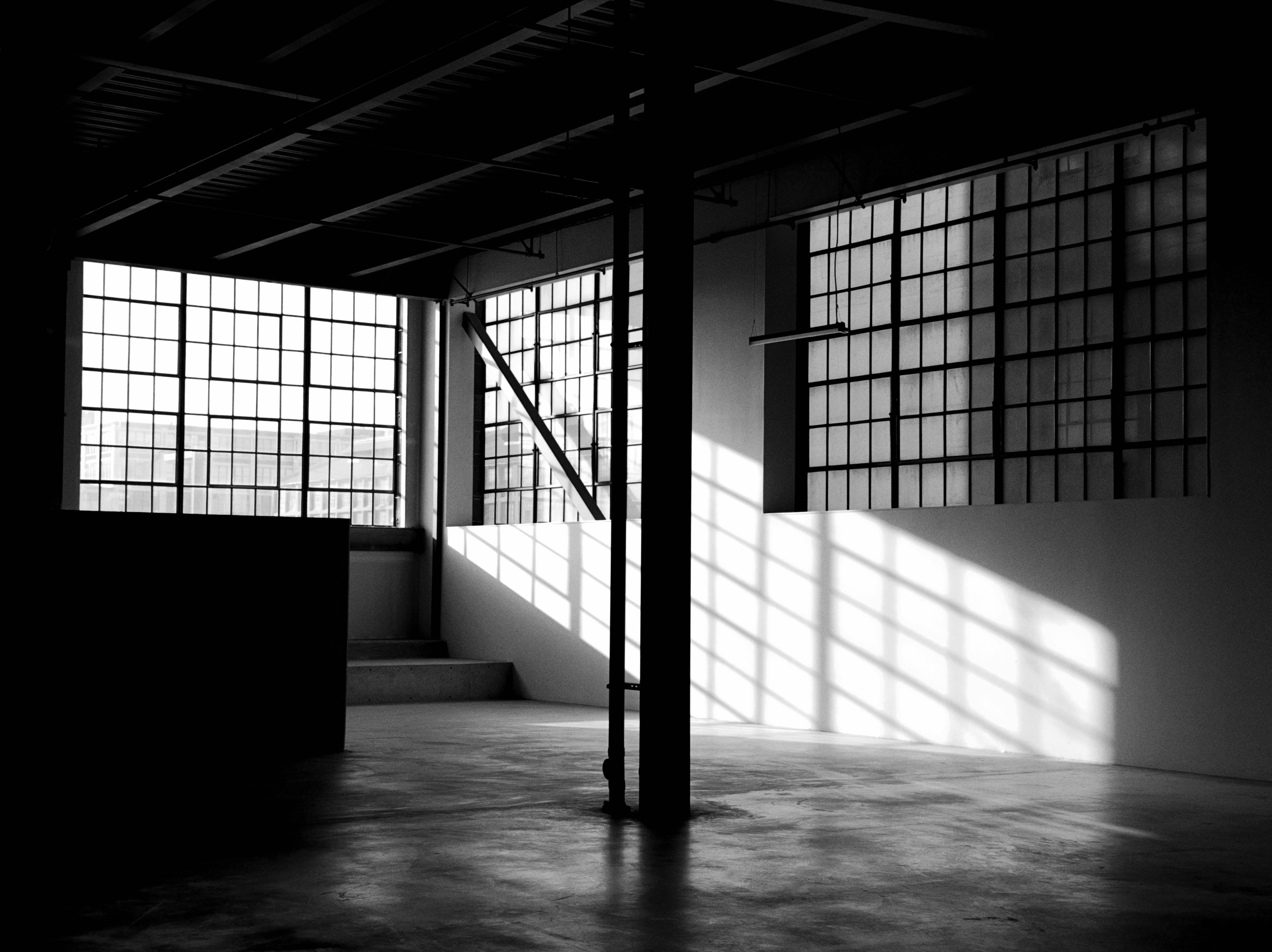 Black and white interior of an empty industrial warehouse with large grid windows and long shadows