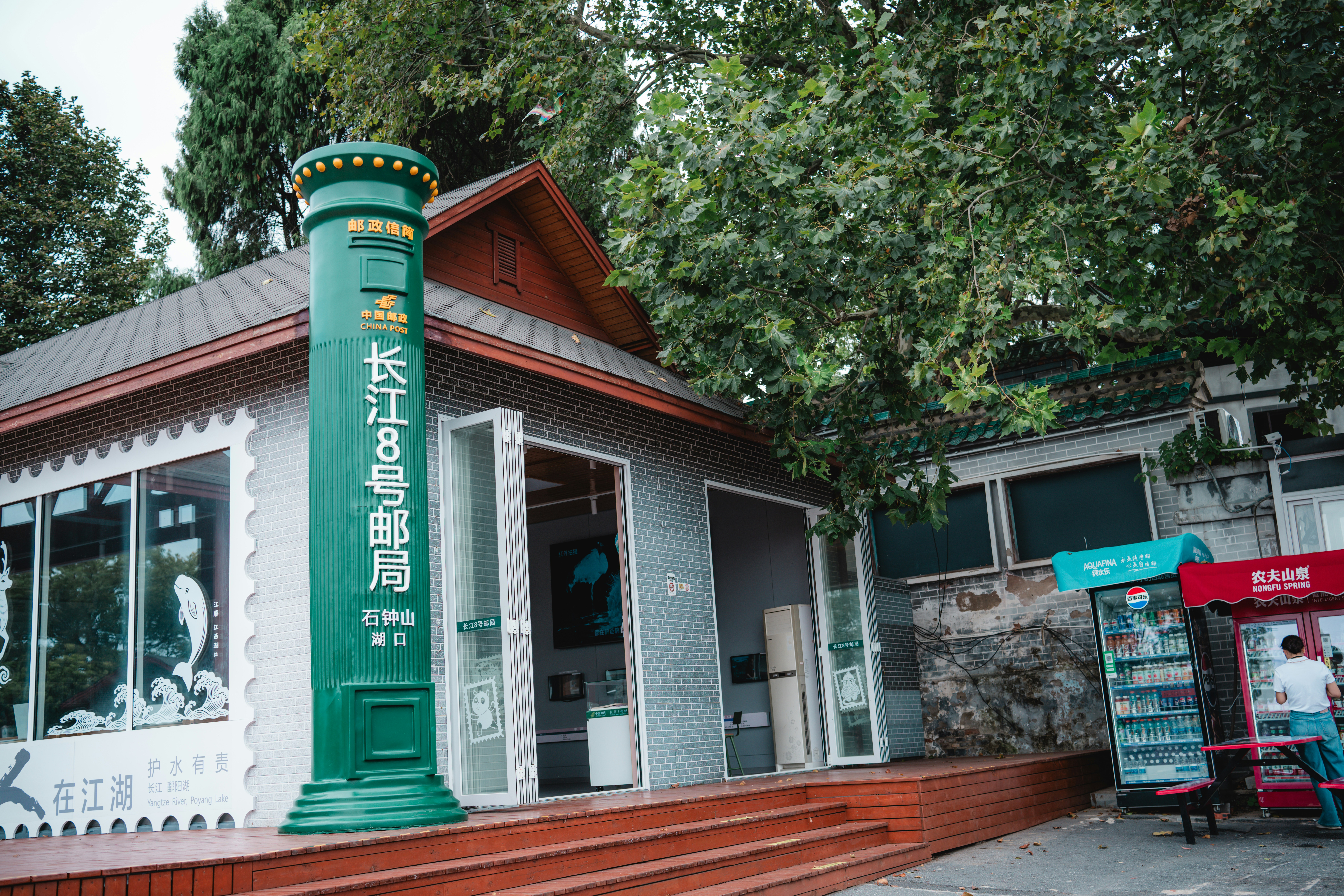 A large green pillar-shaped mailbox outside a grey brick building with a wooden deck