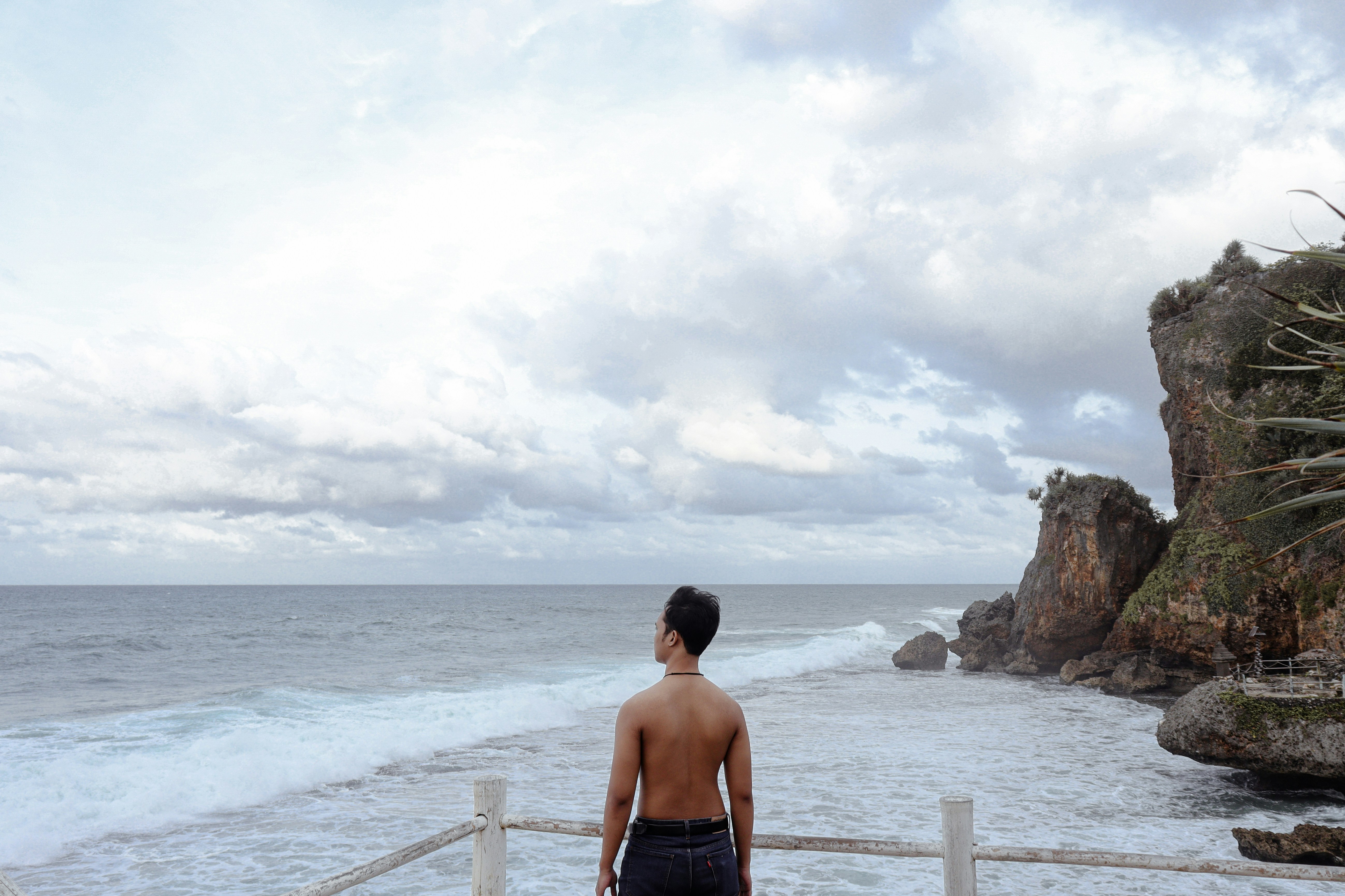 A shirtless man looking out at the ocean from a wooden railing on a cloudy day
