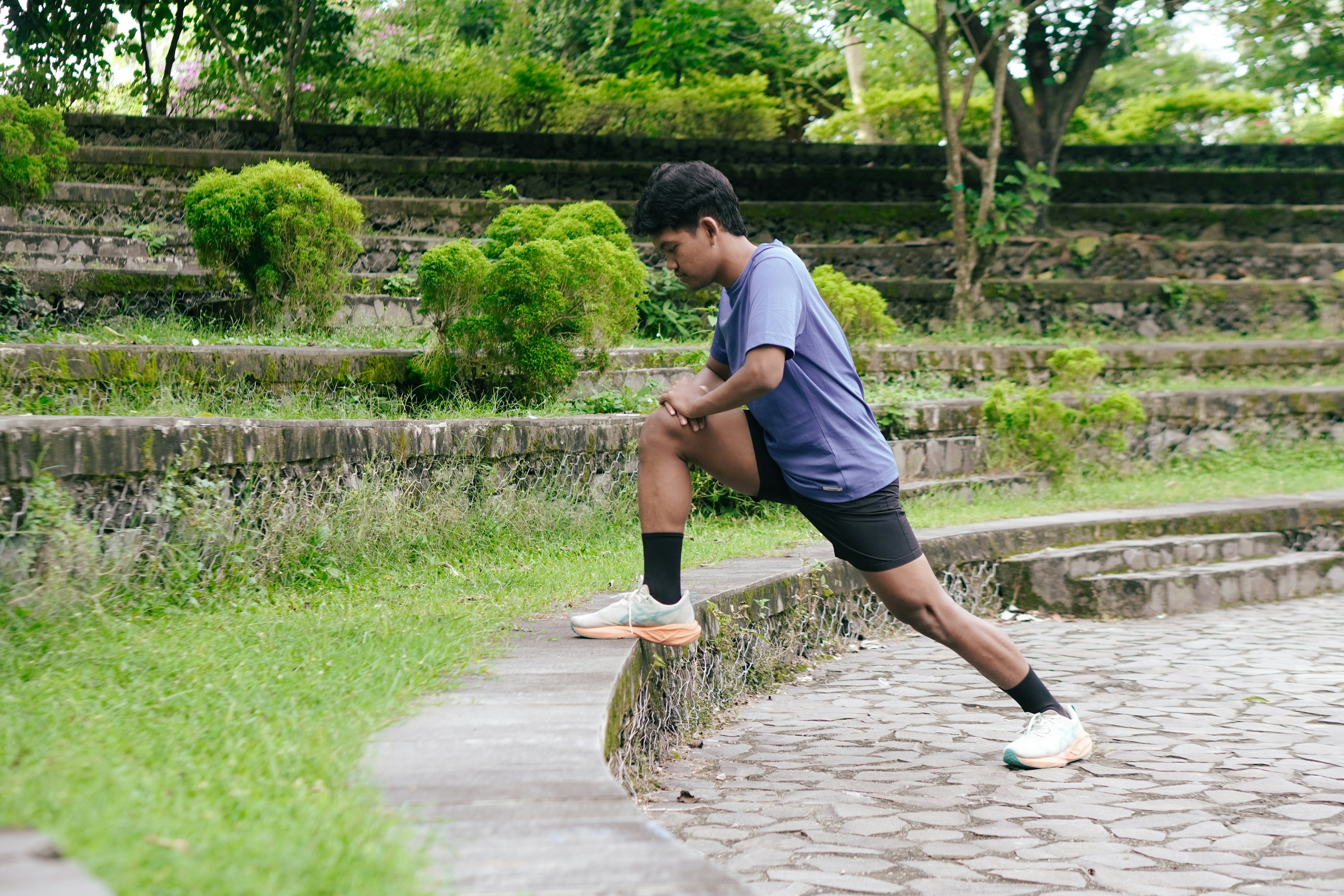 A man in a blue t-shirt stretching his leg on a stone ledge in a park