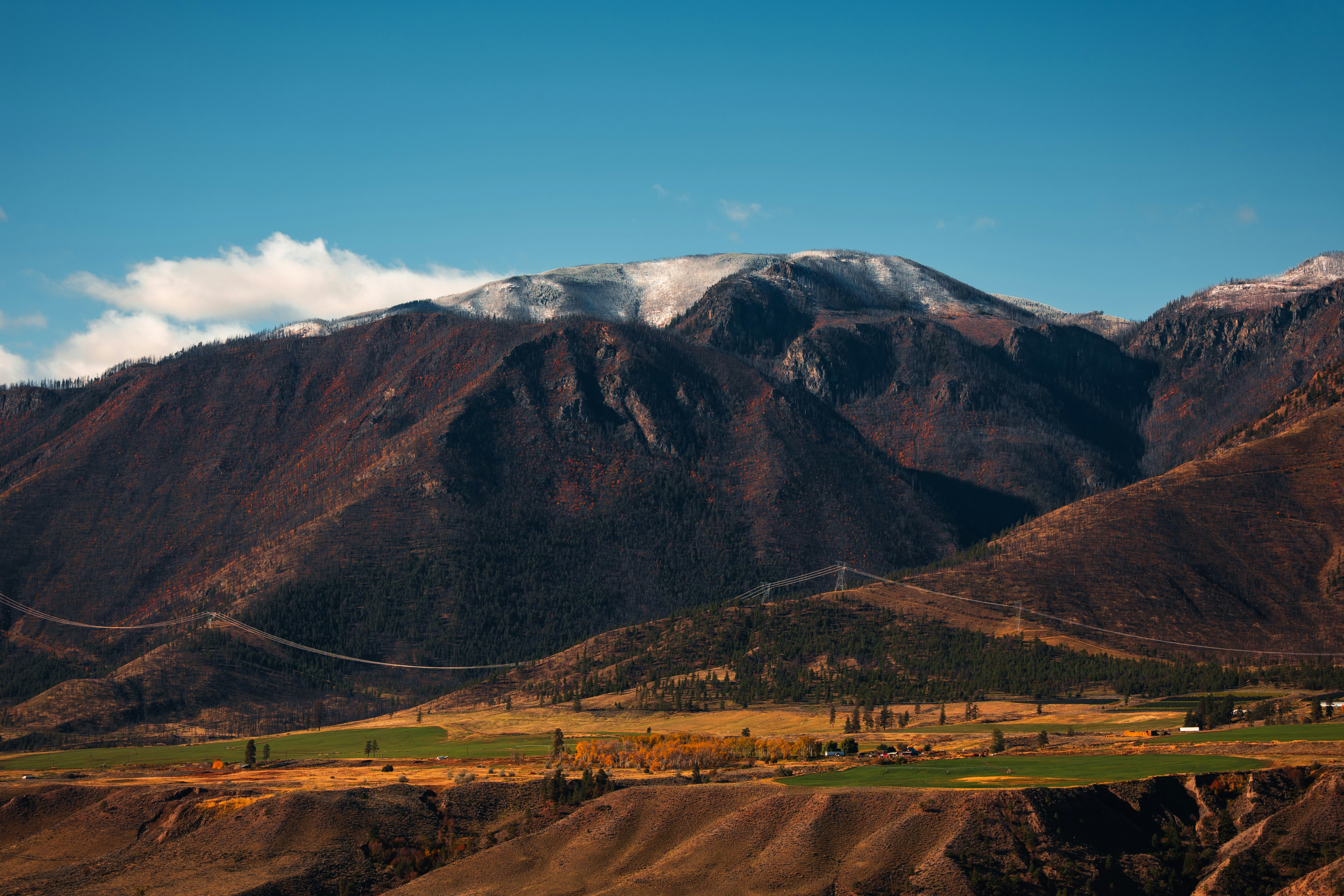 A snow-dusted mountain range rising above a valley with green fields and power lines