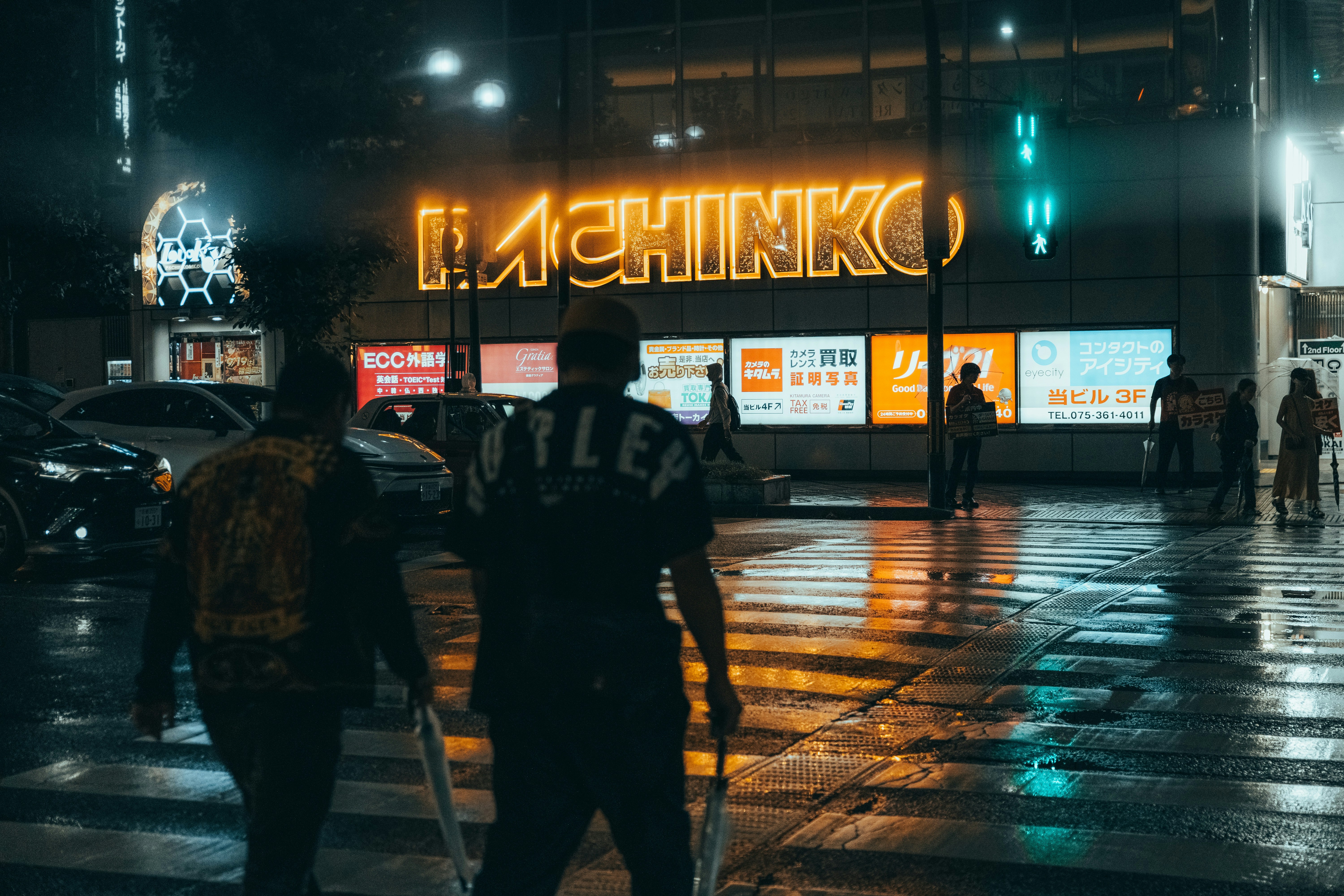 People crossing a wet street at night in front of a glowing neon Pachinko sign