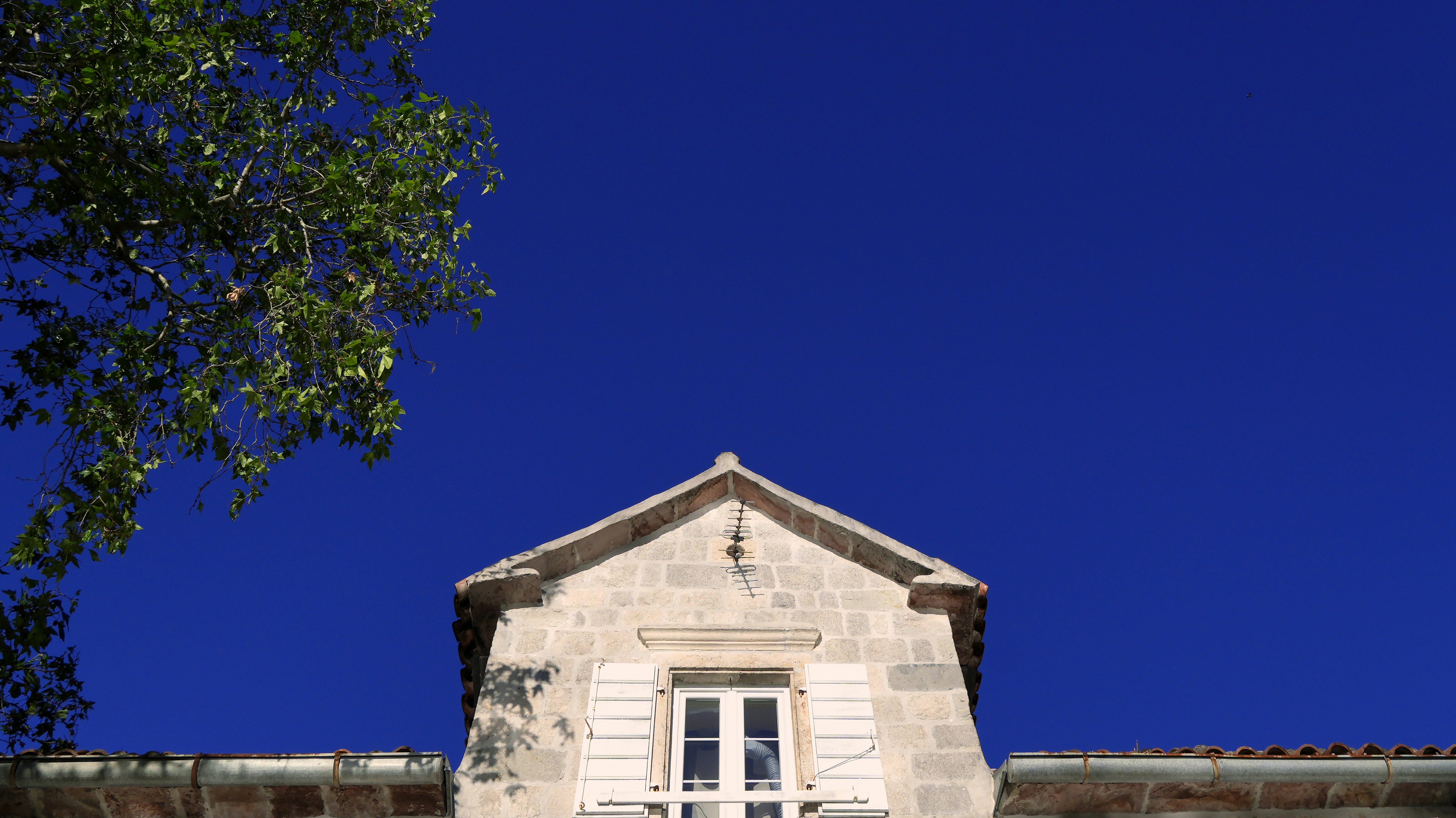 A stone house gable with white shutters under a clear deep blue sky
