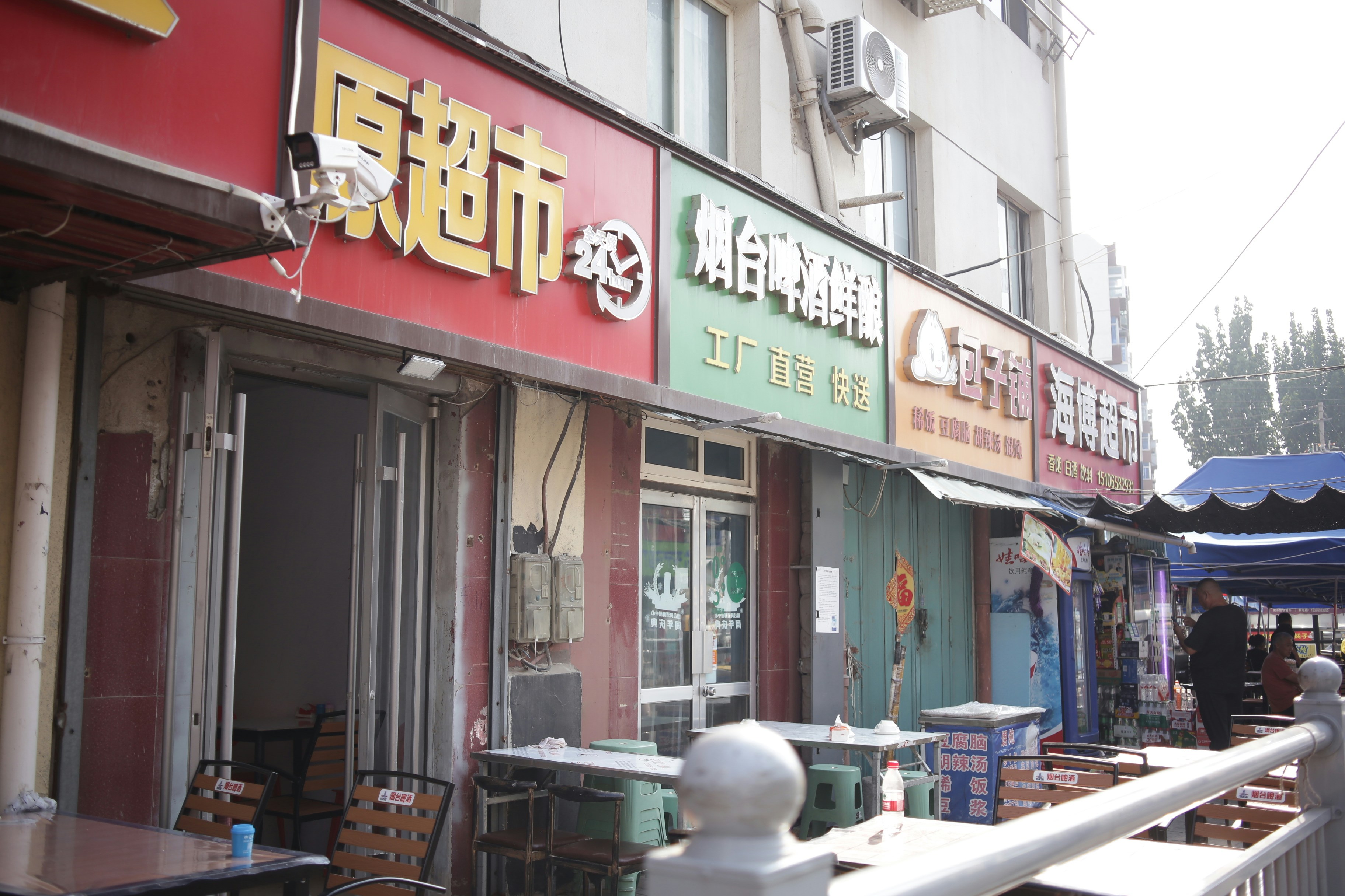 A row of storefronts with colorful signs and outdoor tables on a city street