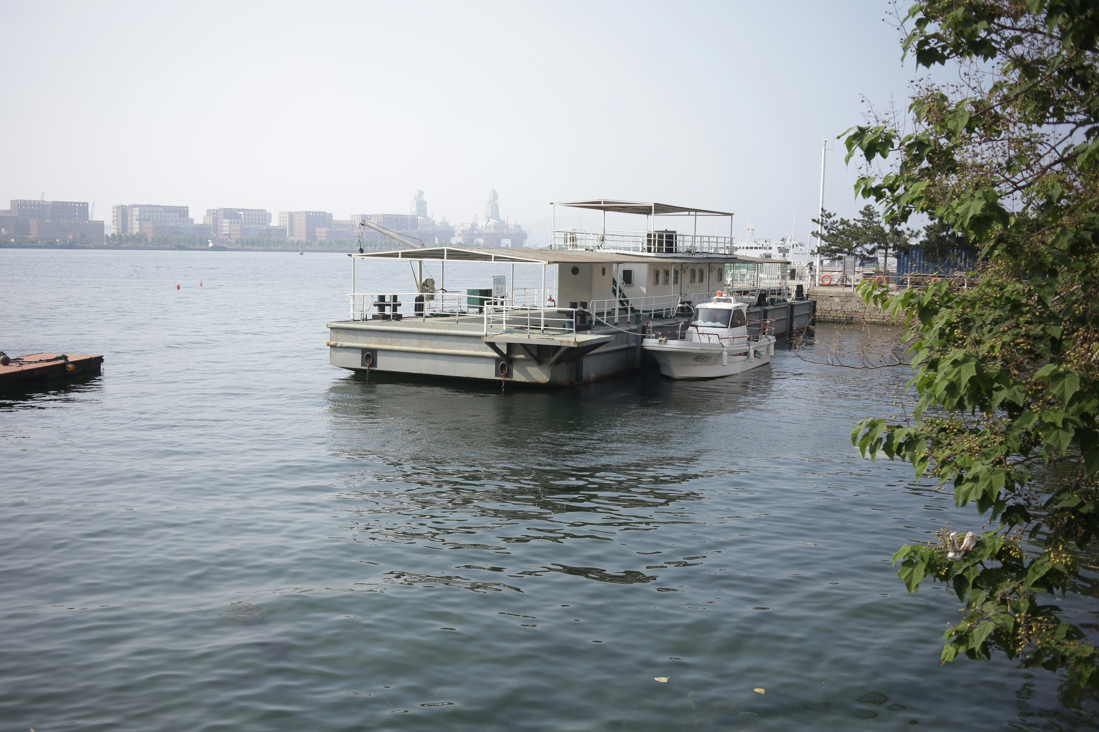 A flat-bottomed barge with a white cabin moored beside a small motorboat in a harbor
