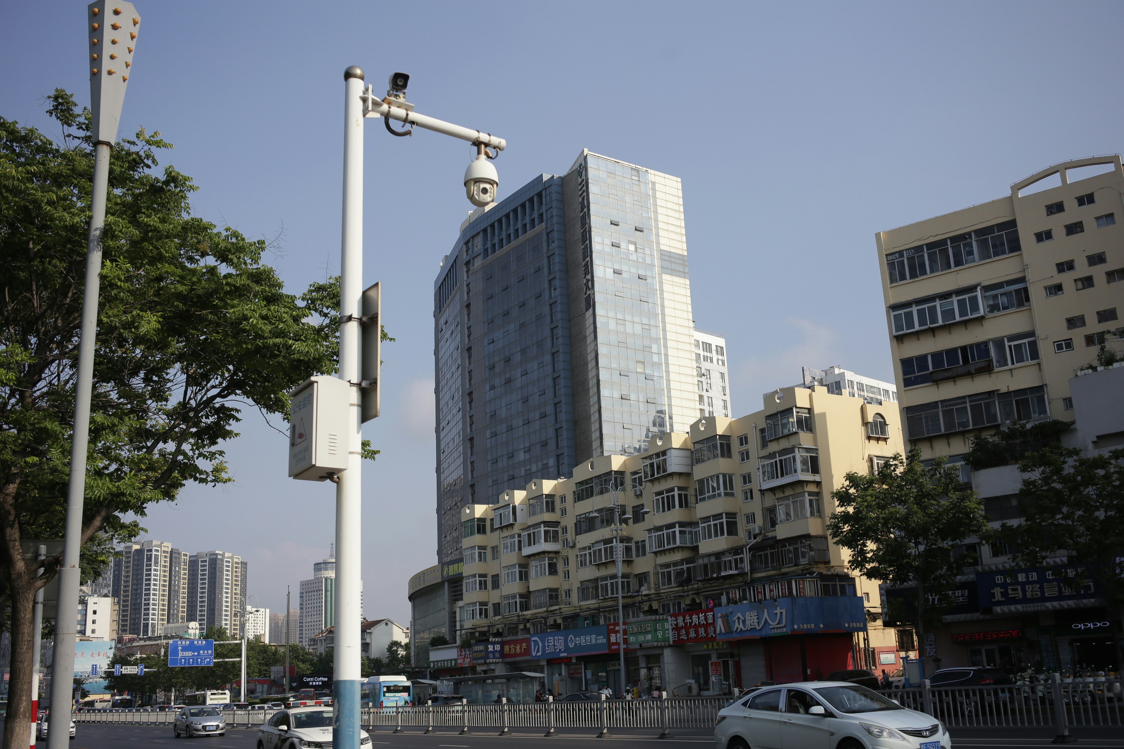 A security camera on a tall pole overlooking a busy city street with apartment buildings
