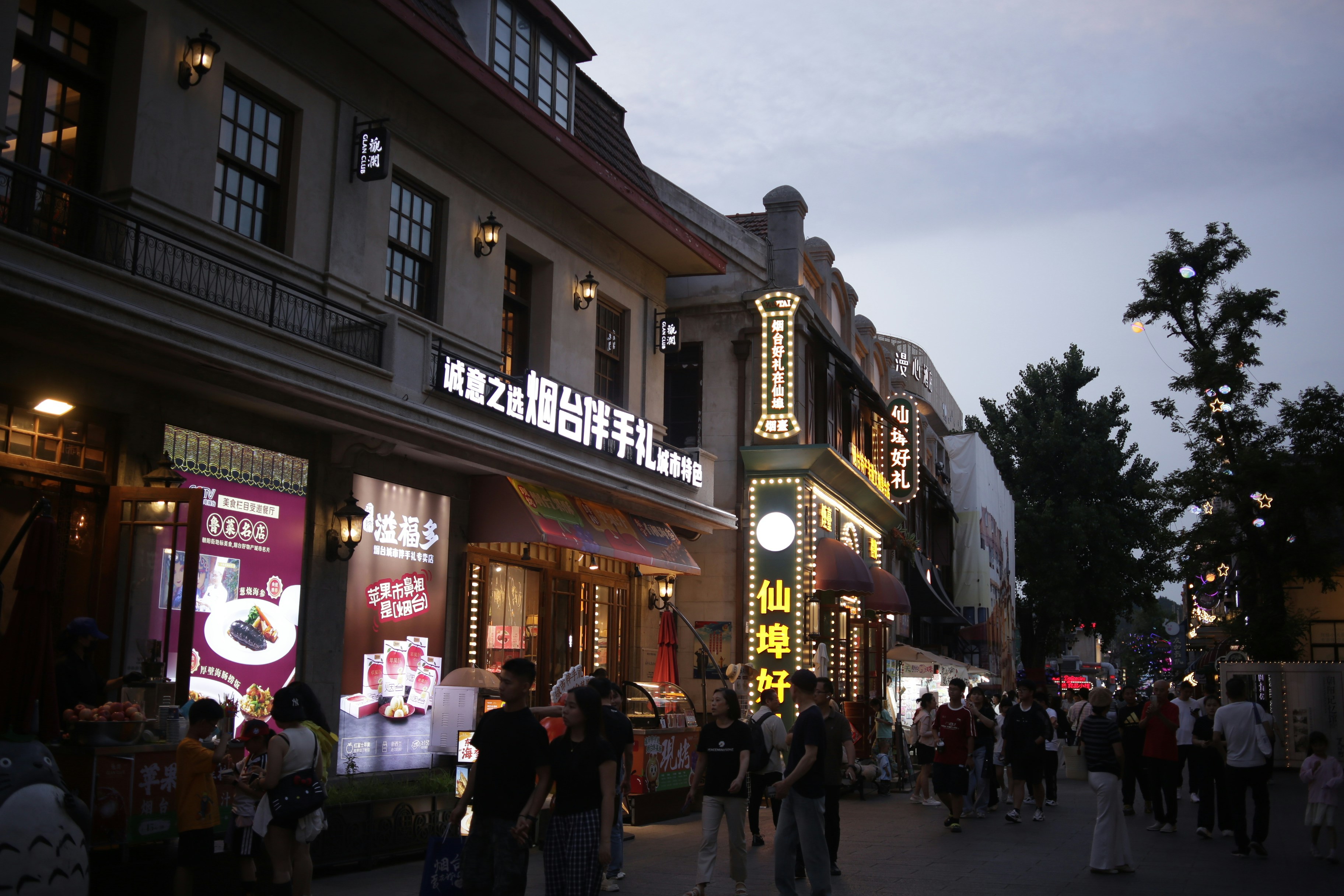 People walking along a pedestrian street lined with illuminated shopfronts and traditional architecture at dusk