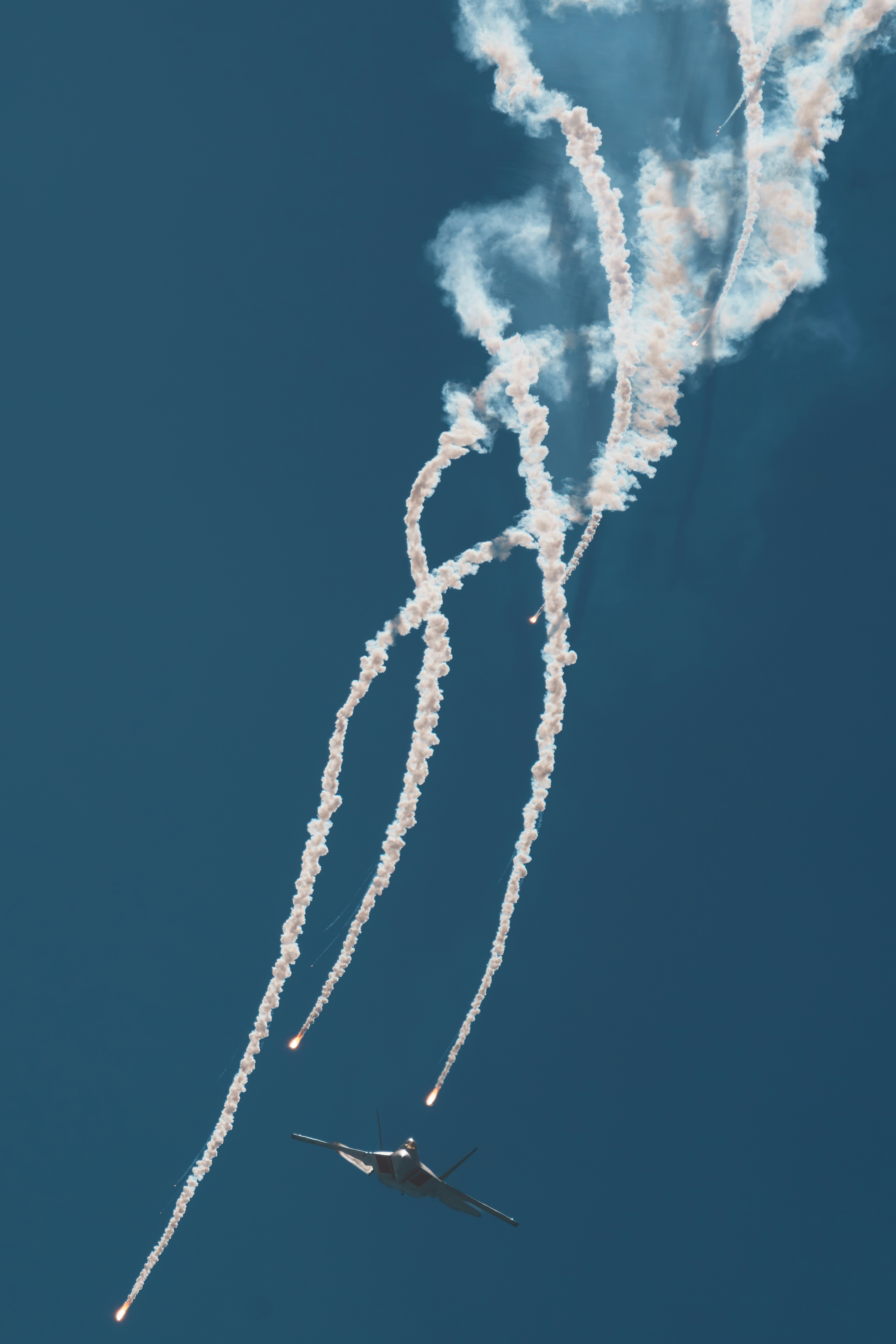 A fighter jet climbing steeply into a clear blue sky while releasing multiple bright flares