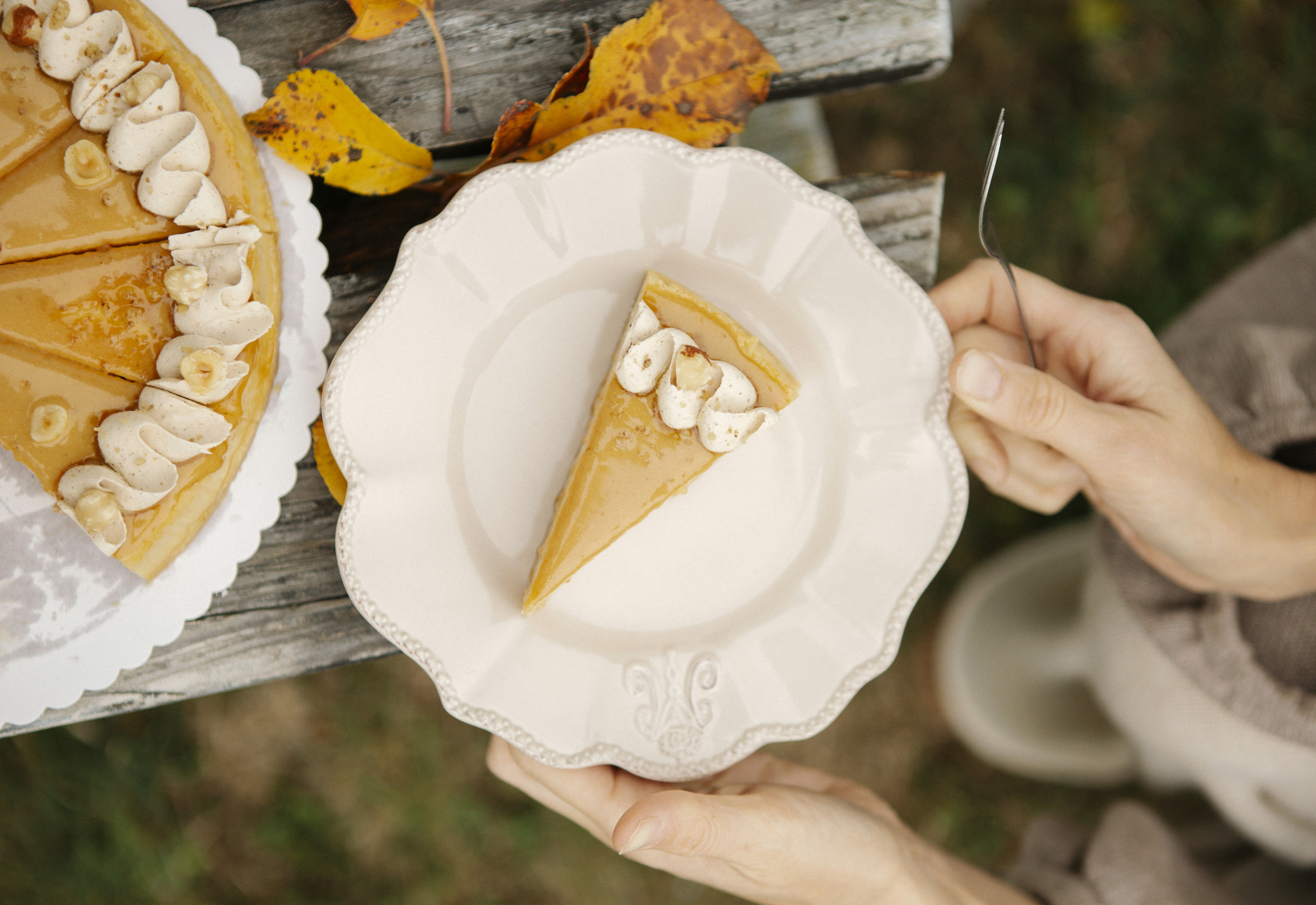 A person holding a plate with a slice of caramel cake topped with whipped cream and hazelnuts