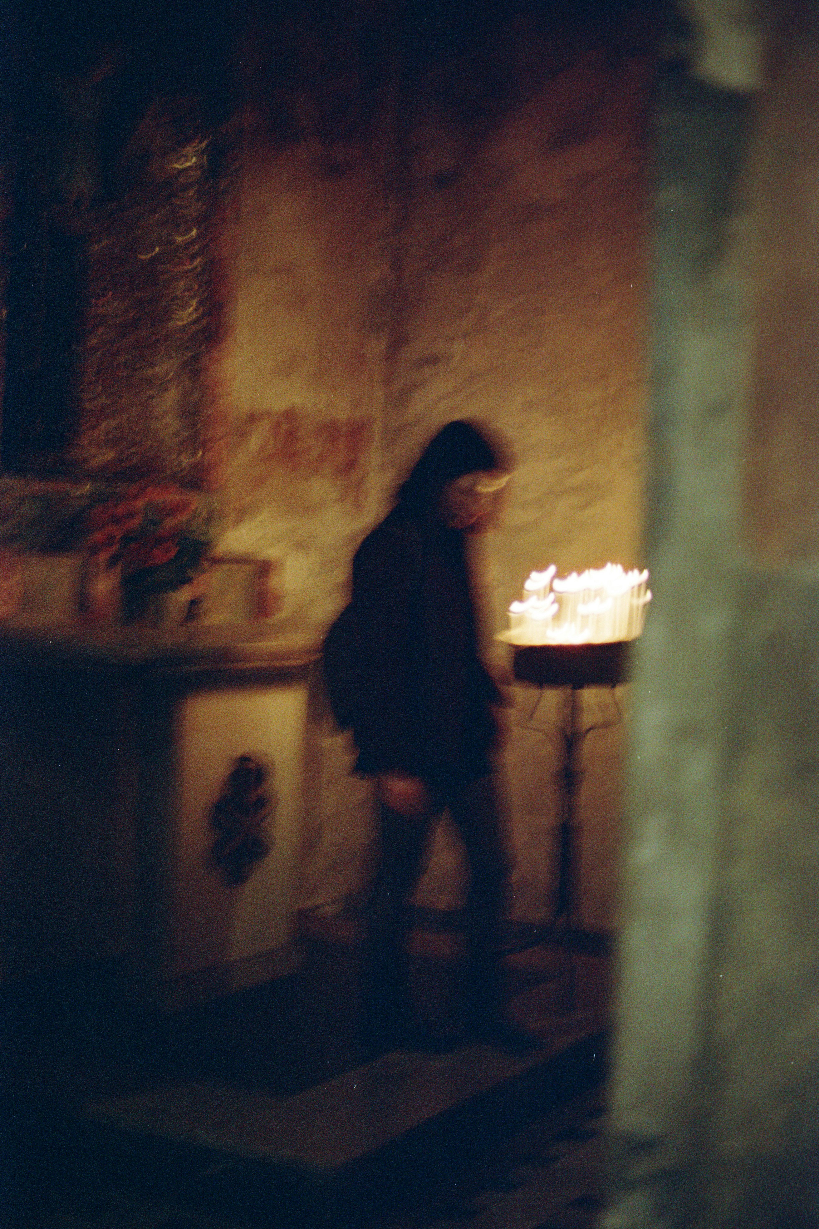 A blurry silhouette of a person walking past a stand of glowing candles in a dim room
