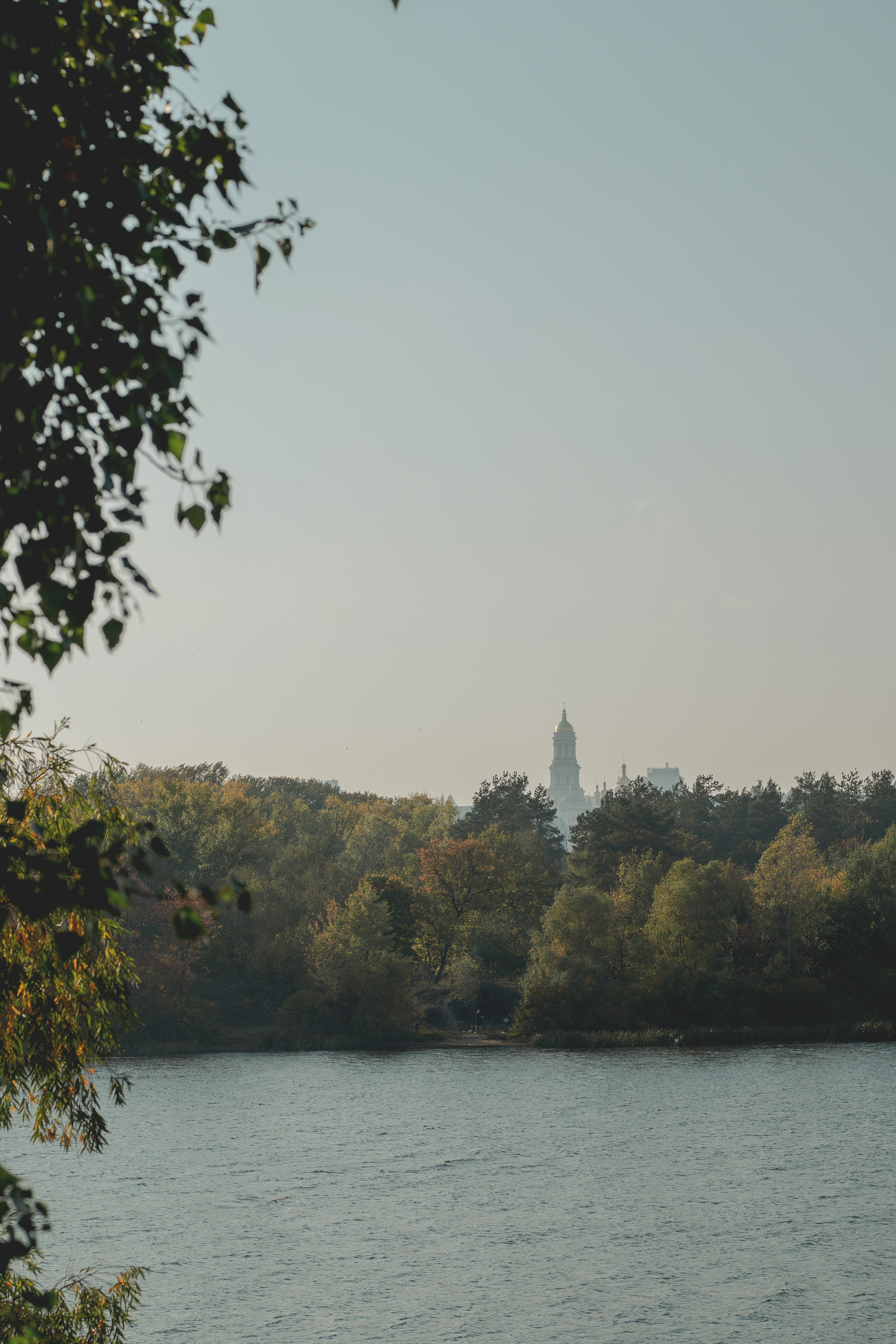 Ein entfernter Kirchturm, der über einem dichten Herbstwald neben einem ruhigen Fluss aufragt