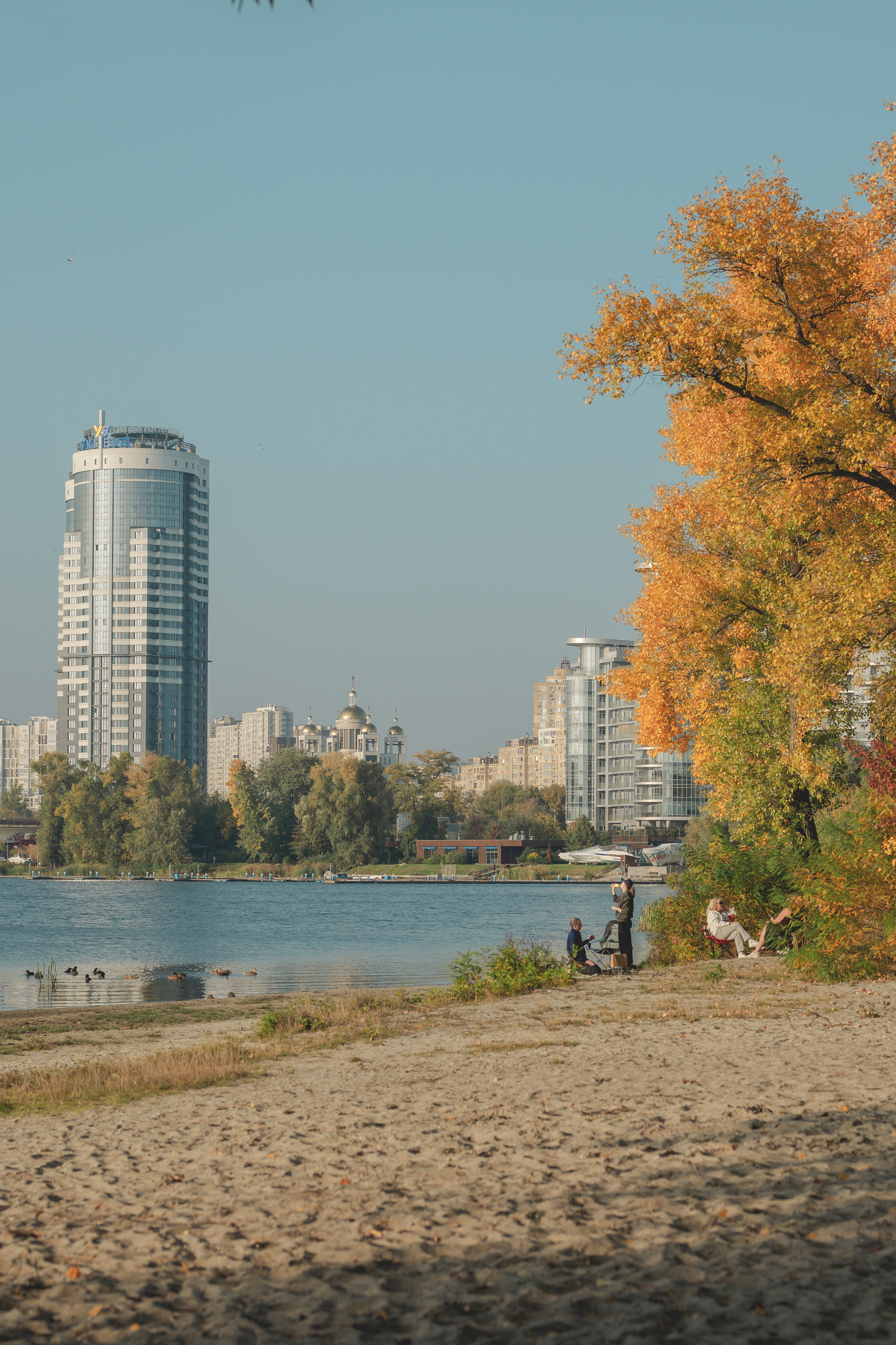 Menschen, die an einem Sandstrand an einem See mit herbstlichen Bäumen und Stadtgebäuden entspannen