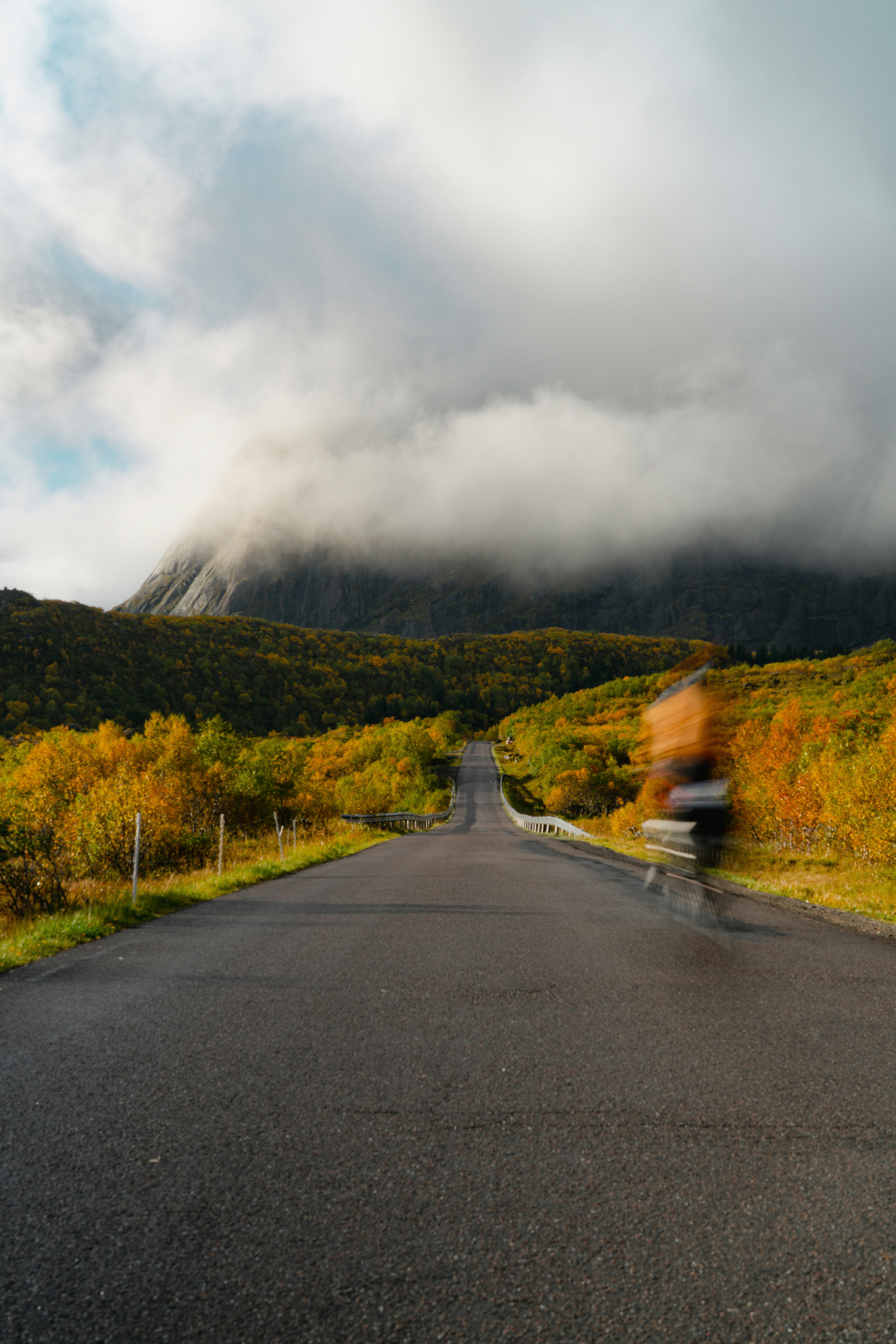 Un ciclista in movimento su una strada attraverso un paesaggio autunnale con una montagna coperta di nuvole