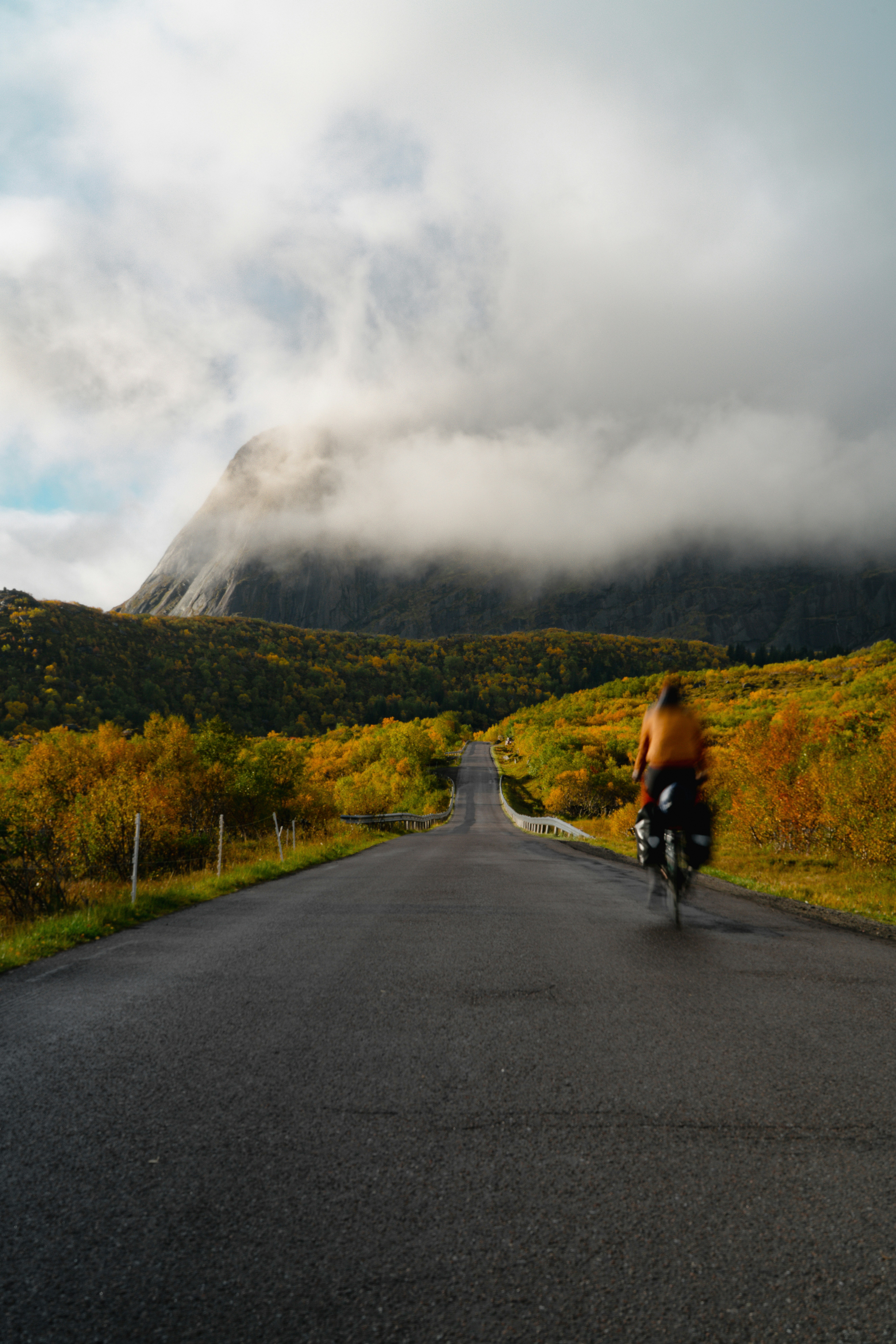 Una persona che pedala su una strada attraverso un paesaggio autunnale verso una montagna coperta di nuvole