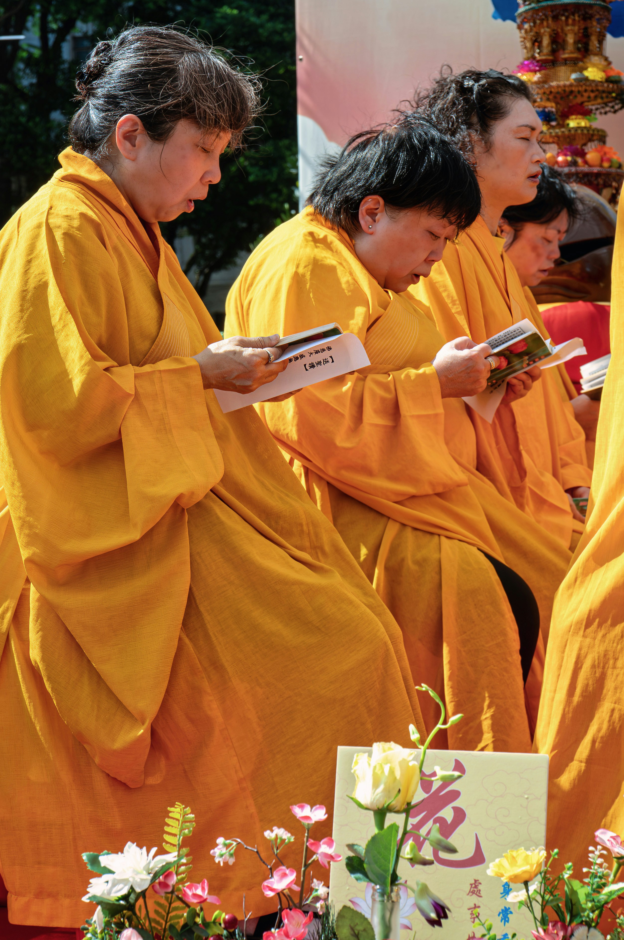 Three people in saffron robes reading from small booklets during an outdoor ceremony