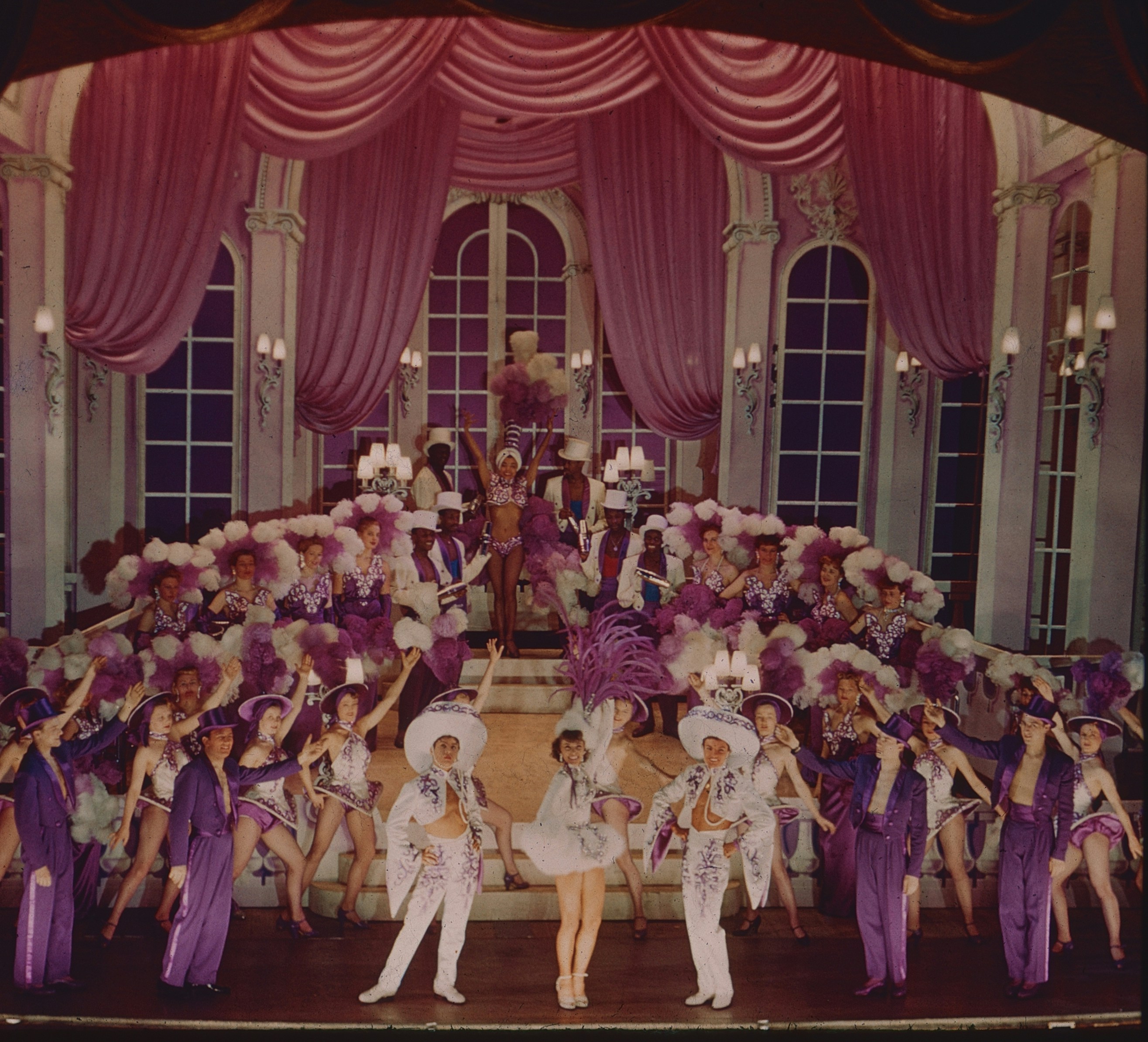 debutant: A large troupe of performers in purple and white costumes on a stage with pink curtains