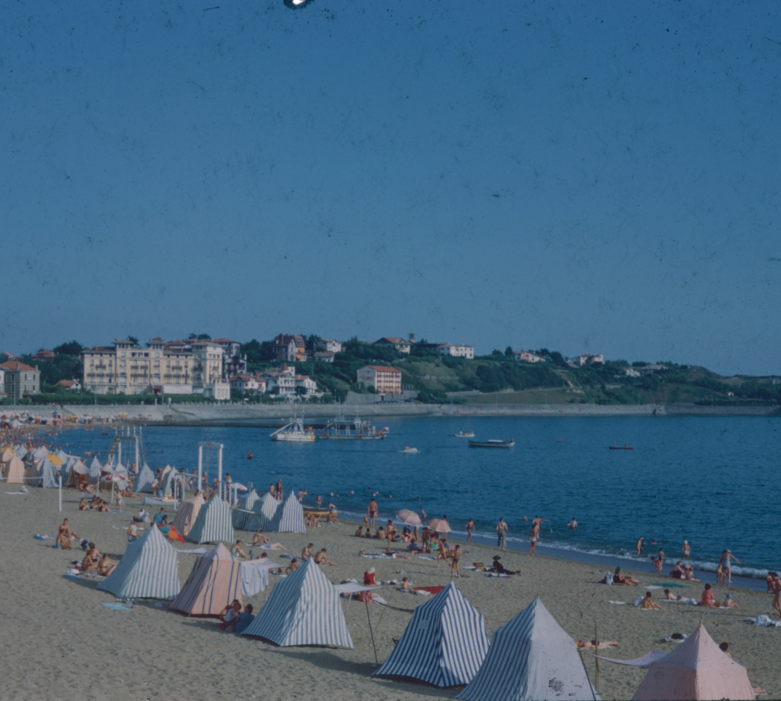 People on a sandy beach with striped tents and a coastal town in the distance