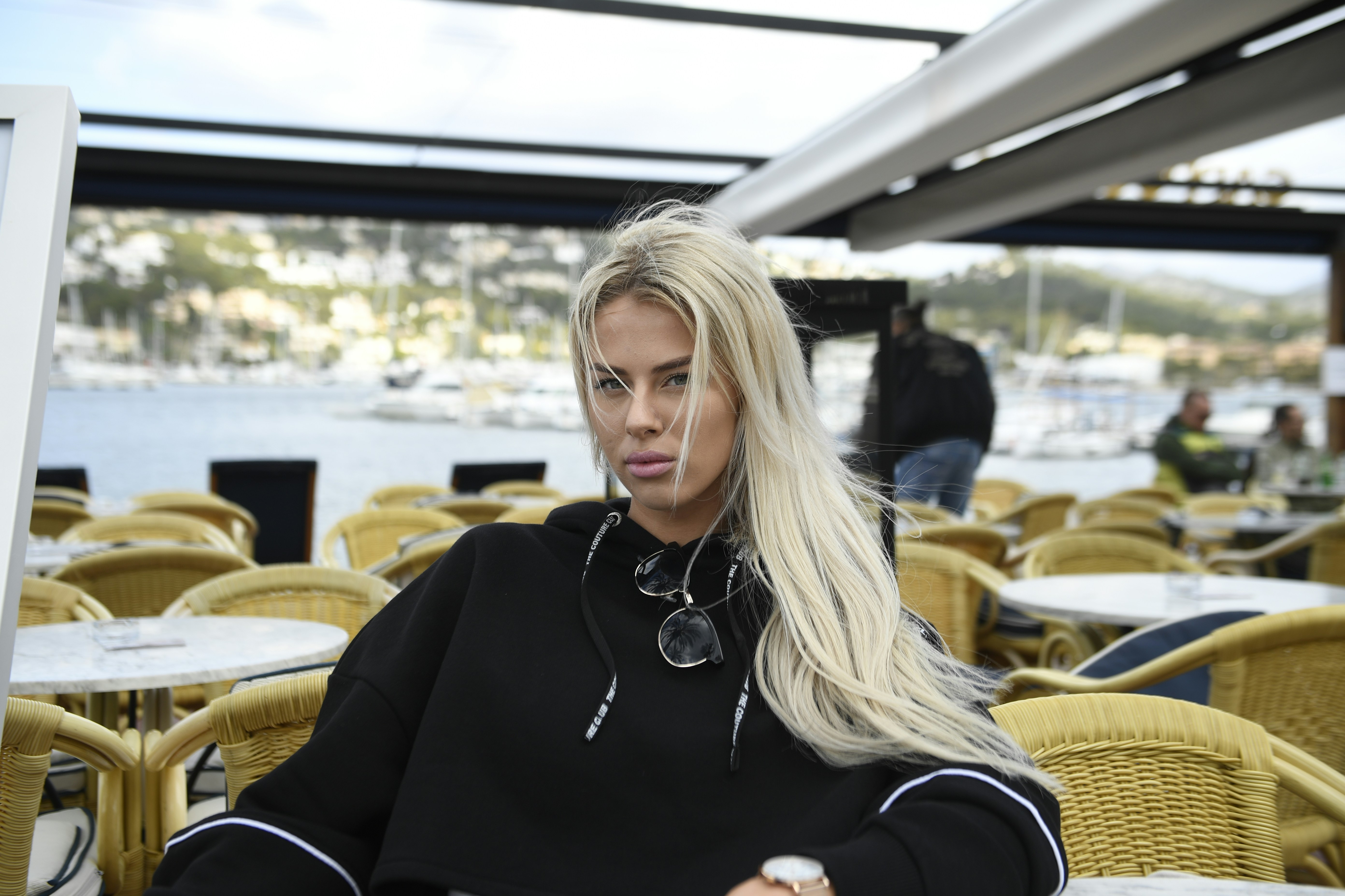 A blonde woman in a black hoodie sitting at a cafe table by a harbor
