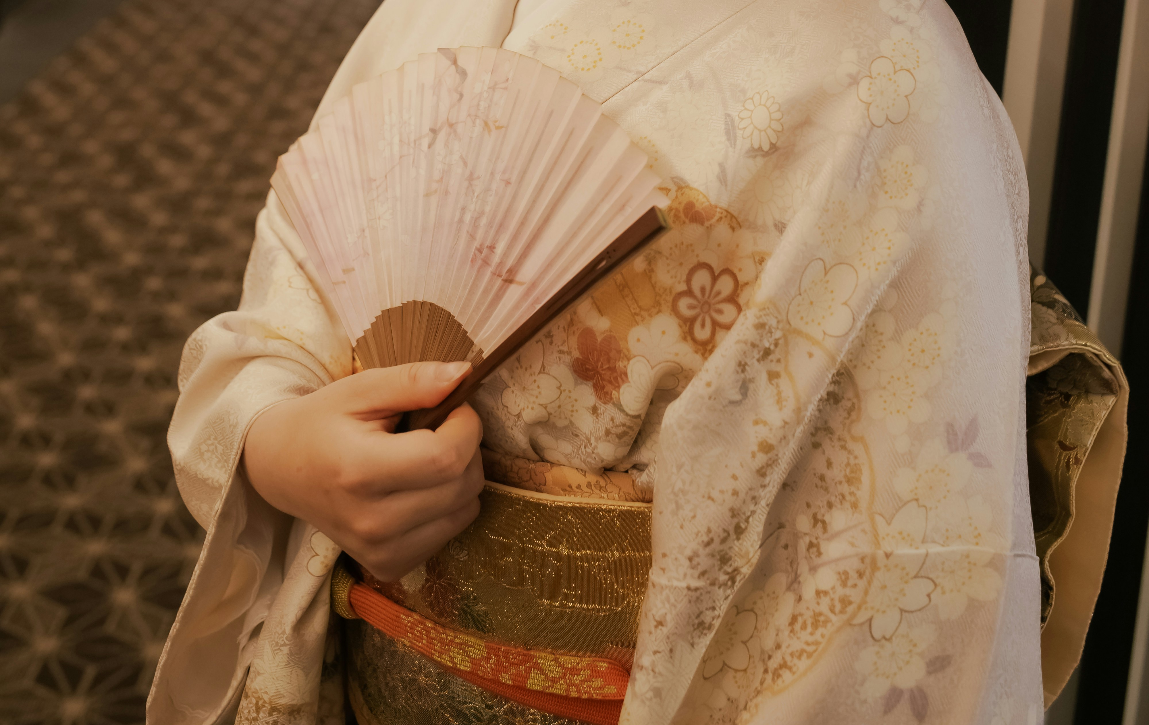 A person wearing a cream kimono with floral patterns holding a folded pink fan