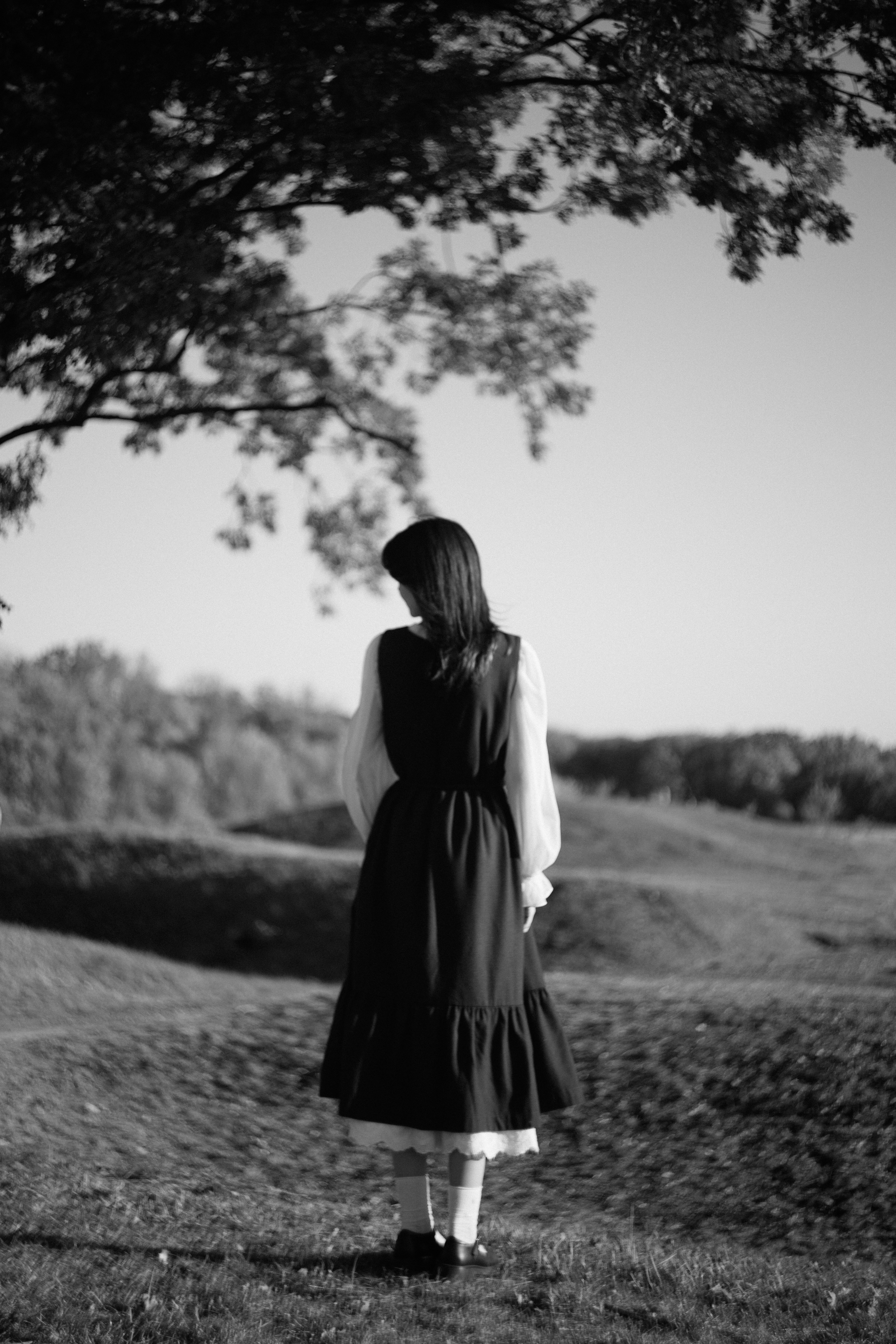 A woman in a dark dress and white socks standing in a field under a tree