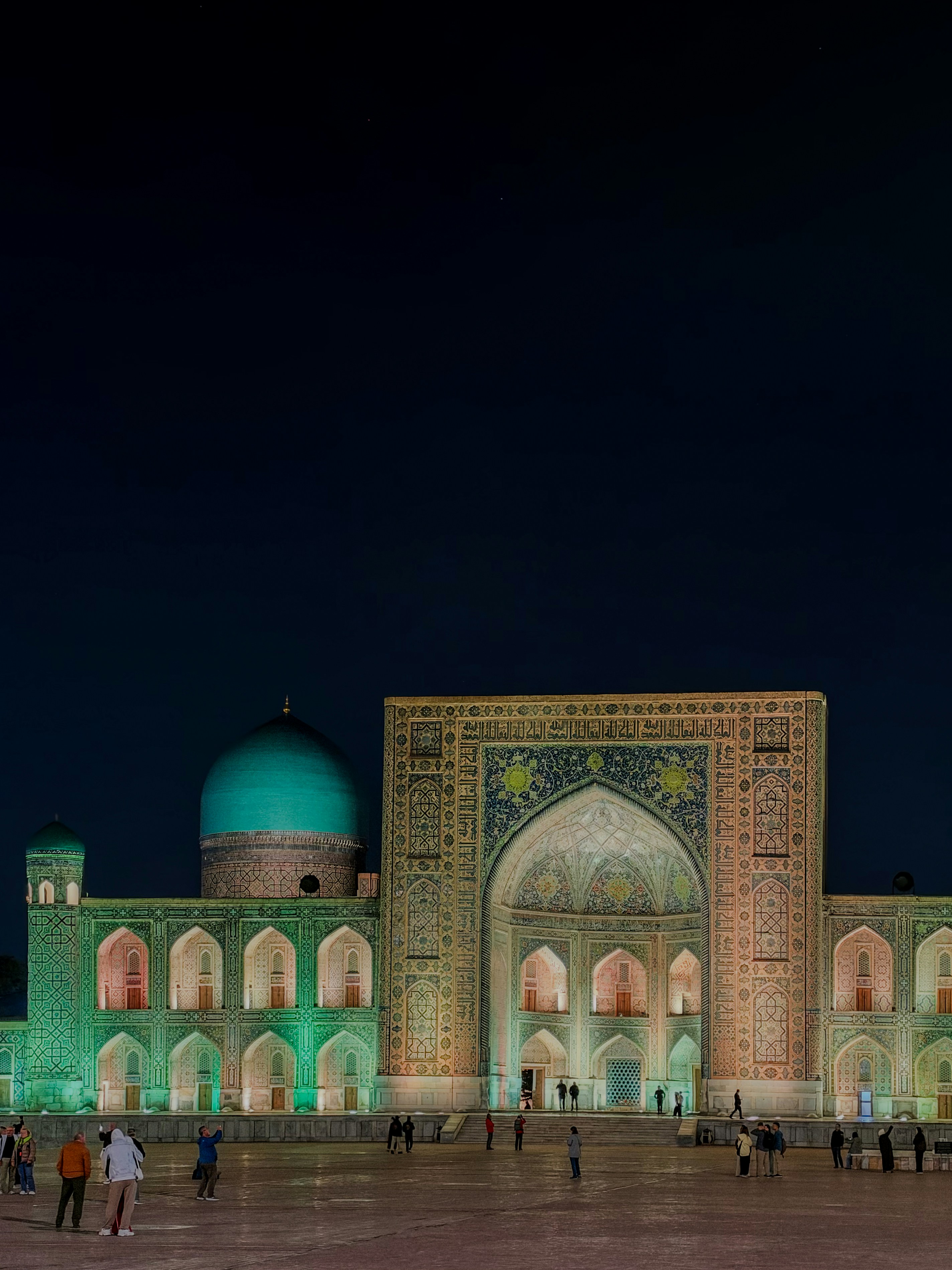 The illuminated Tilya-Kori Madrasah at night with people walking in the Registan square in Samarkand