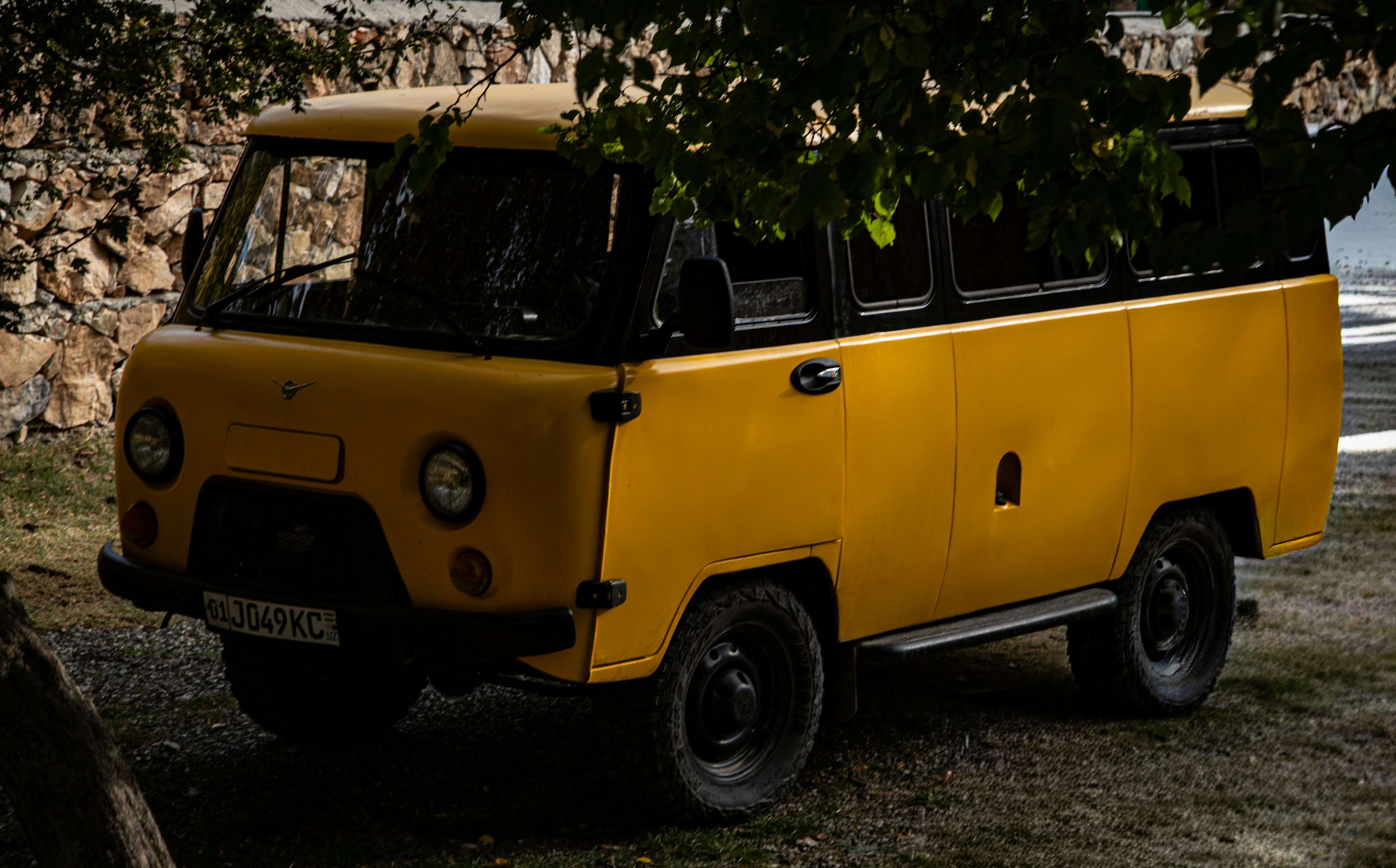A yellow UAZ Bukhanka van parked on a gravel surface beneath tree branches