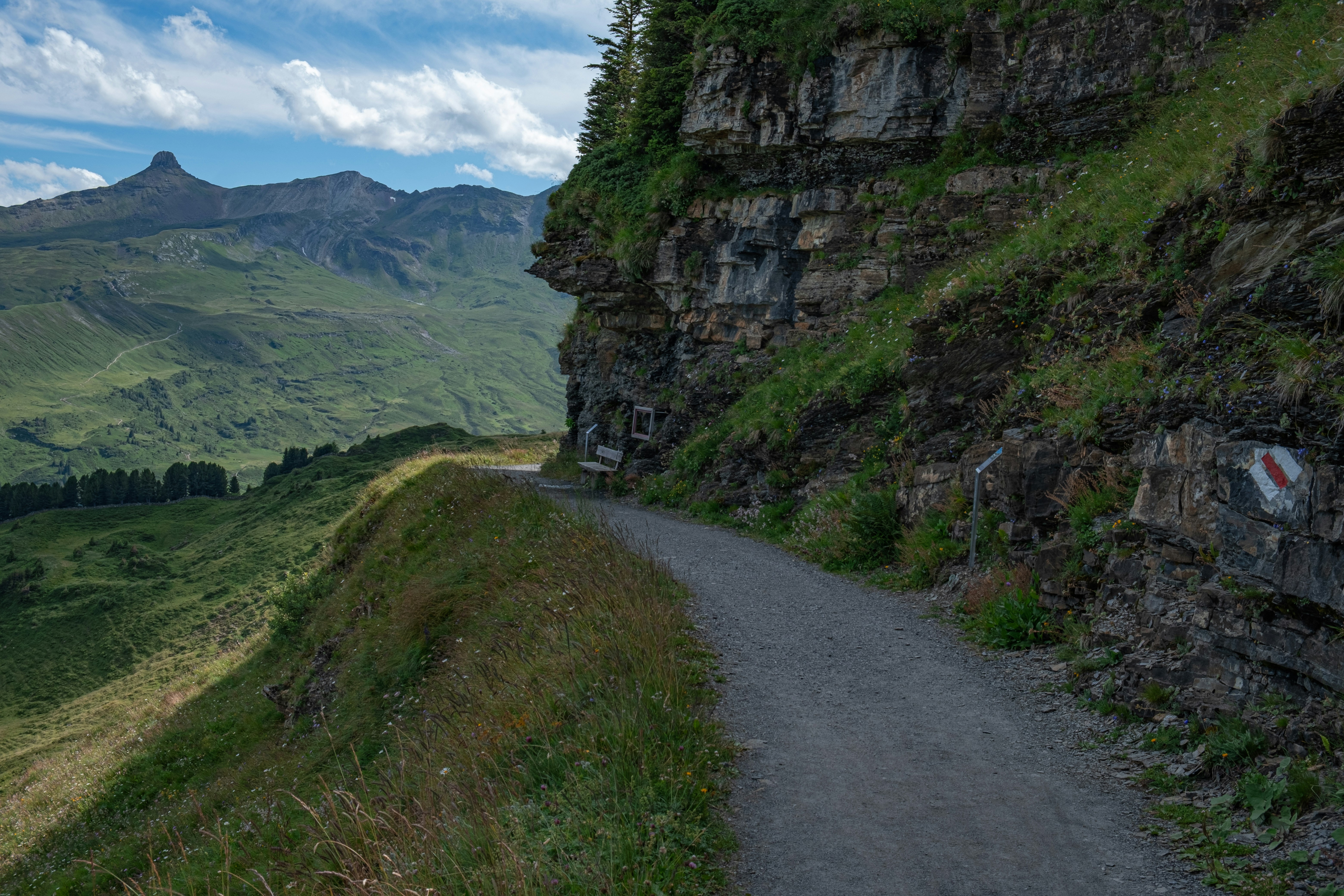 Ein Wanderweg entlang einer Bergklippe mit Blick auf ein grünes Tal mit entfernten Gipfeln unter einem teilweise bewölkten Himmel.