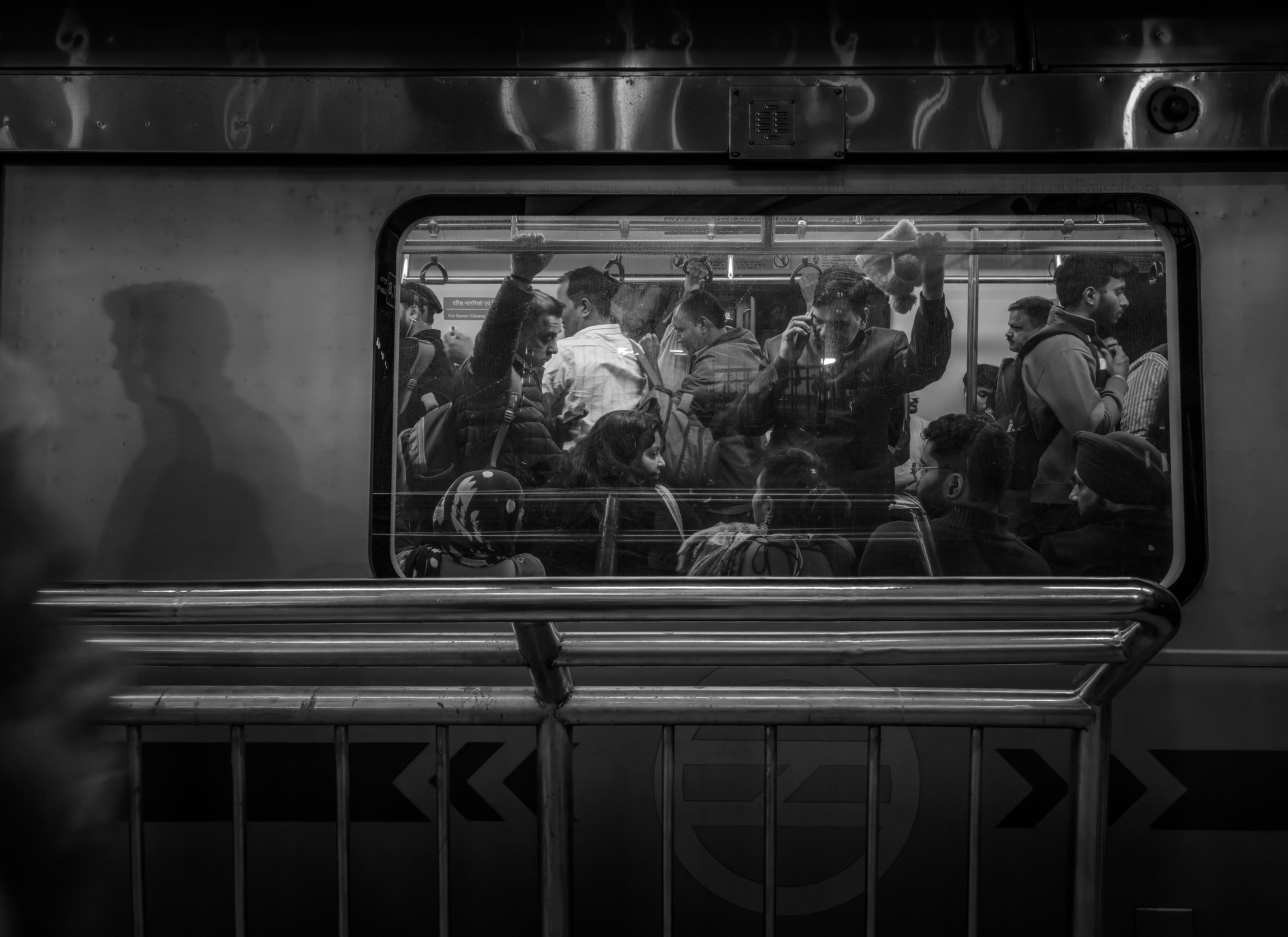 A black and white view through a Delhi metro train window at crowded passengers